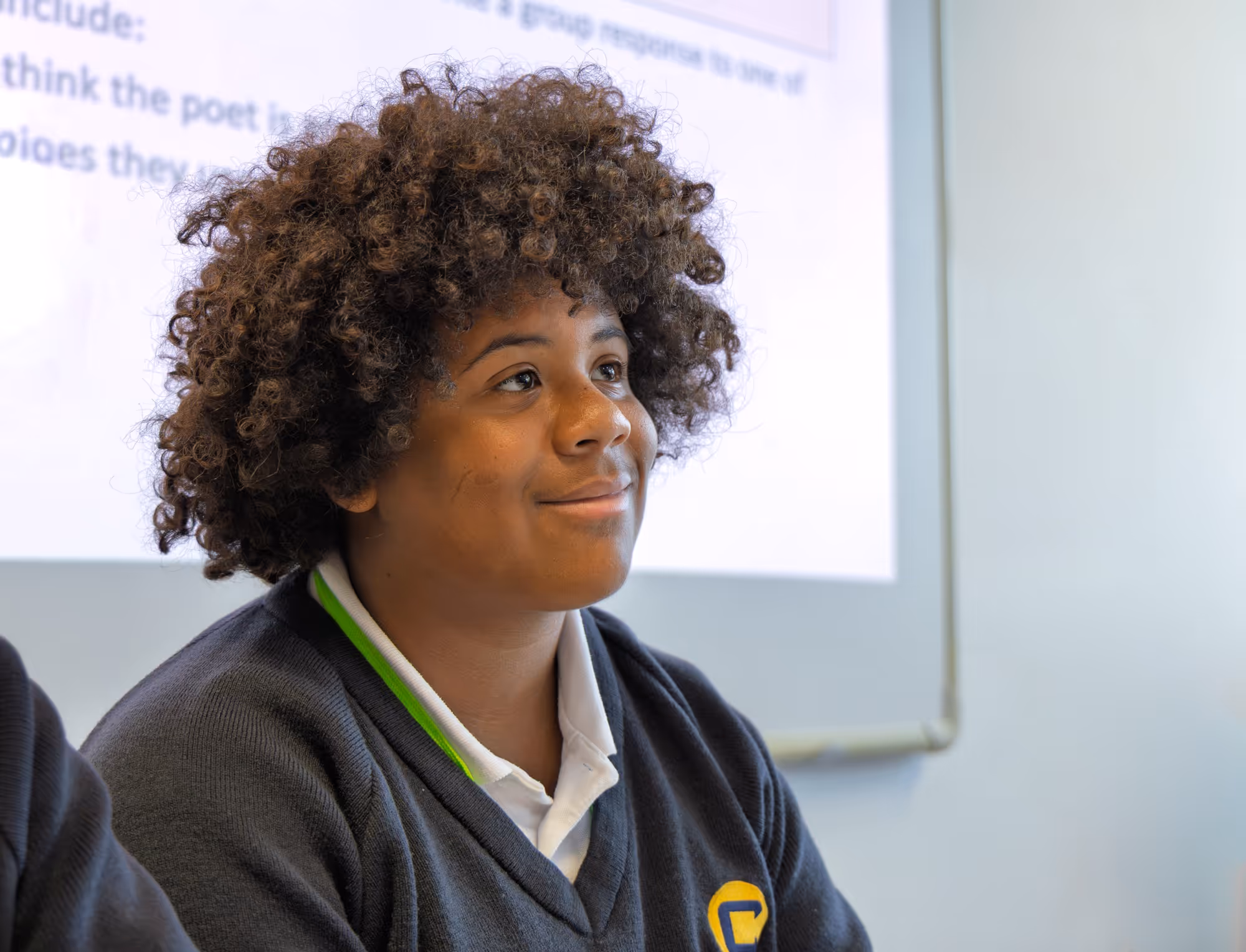 Smiling student with curly hair wearing a dark school uniform sitting in a classroom with a whiteboard in the background.