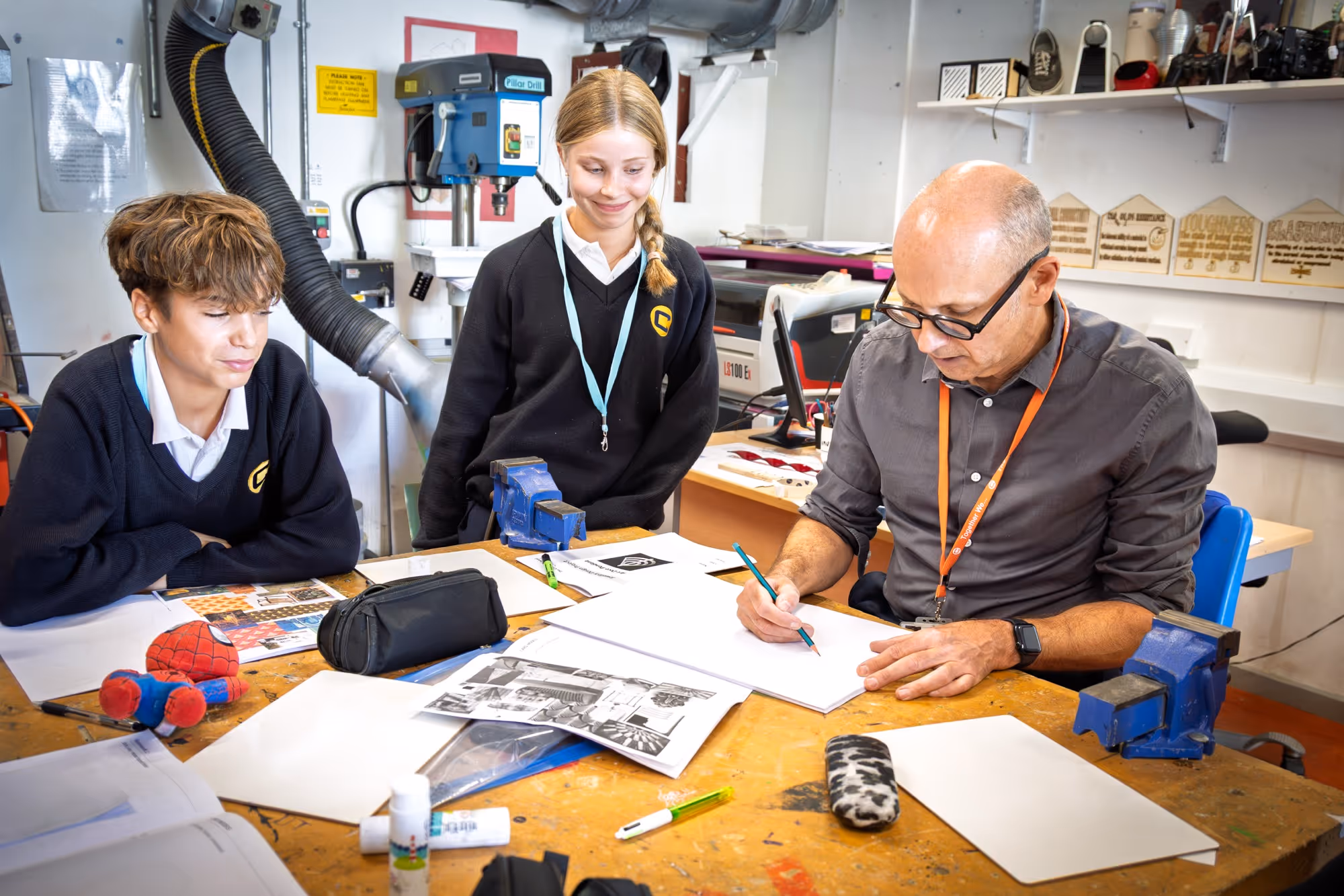 A man with glasses and two students sitting at a cluttered workshop table with papers, pencils, and a Spider-Man toy; the man is drawing while the students watch attentively.