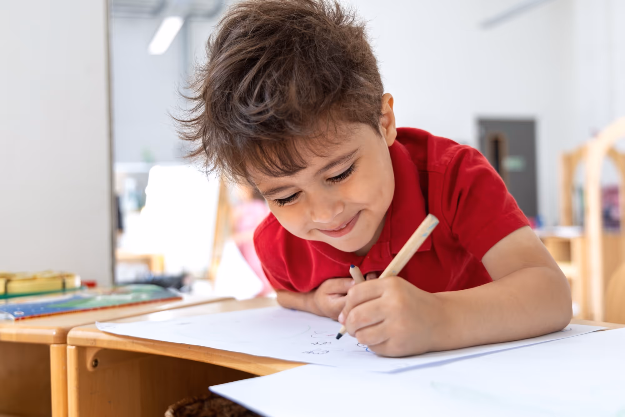Young boy with messy brown hair wearing a red shirt smiling as he writes on white paper at a wooden desk in a bright classroom.