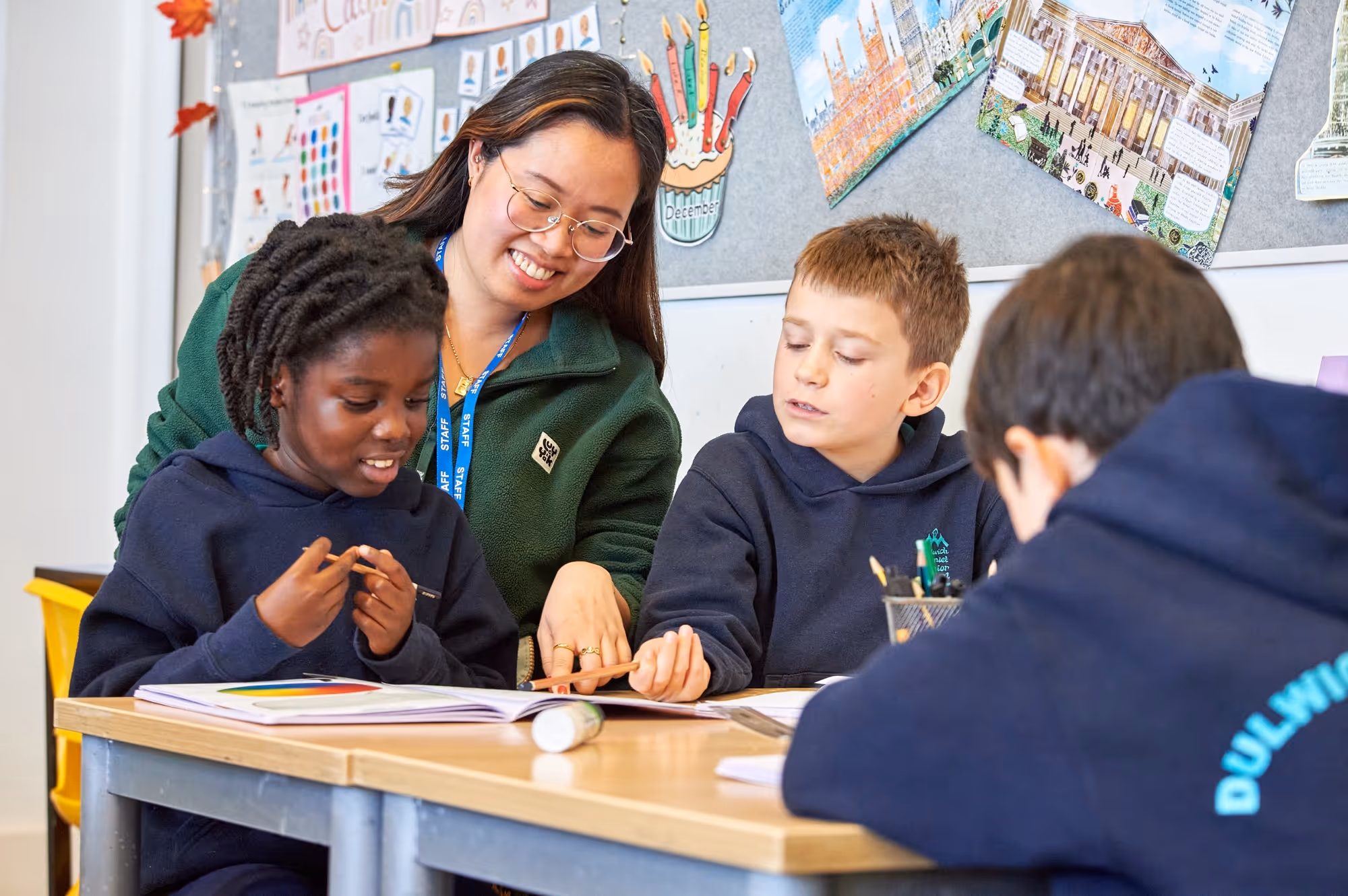 A female teacher with glasses helps two elementary students sitting at a desk while another student works nearby in a classroom.