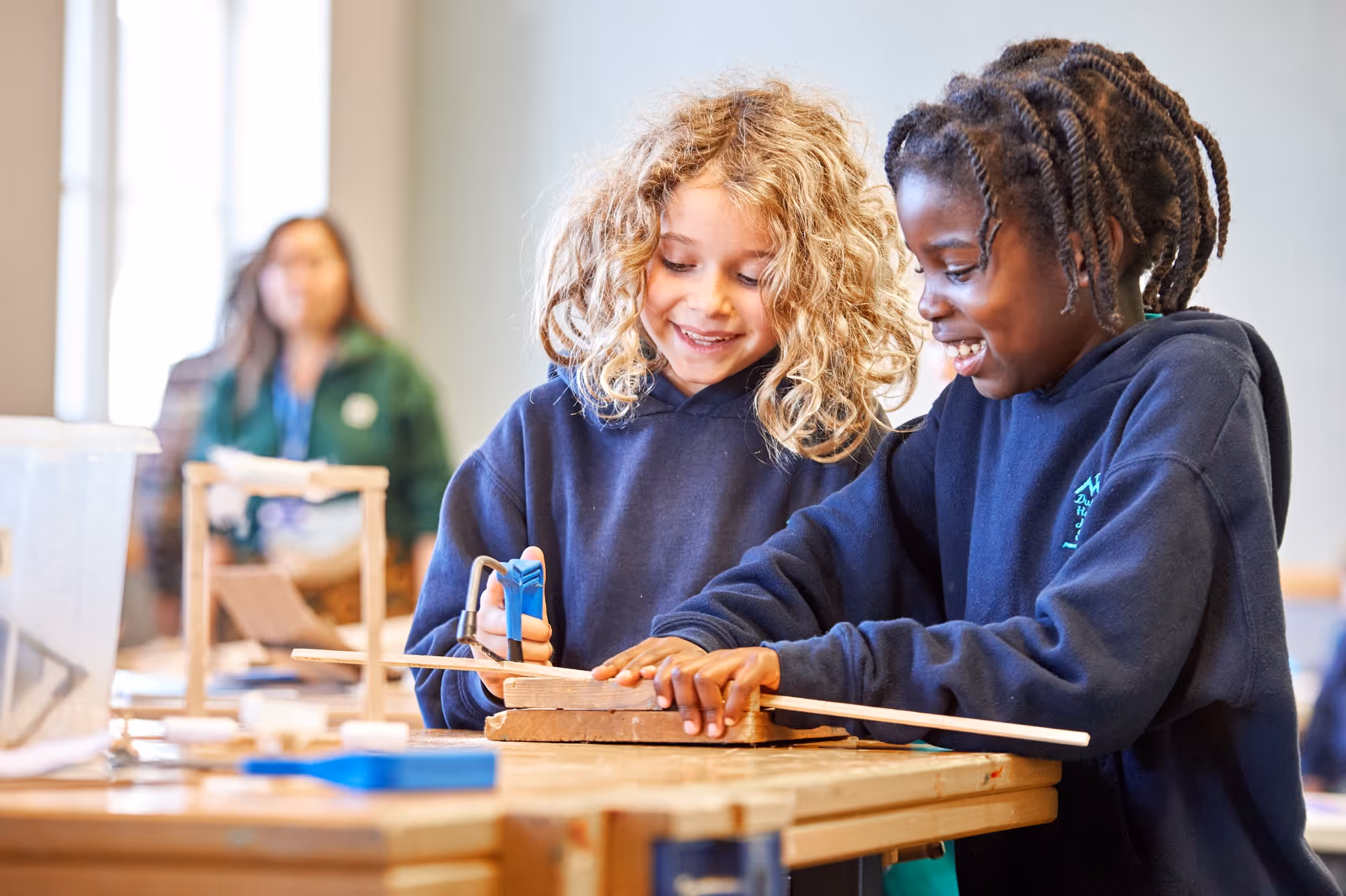 Two children smiling and working together on a woodworking project with a handsaw and wooden pieces on a table in a classroom setting.