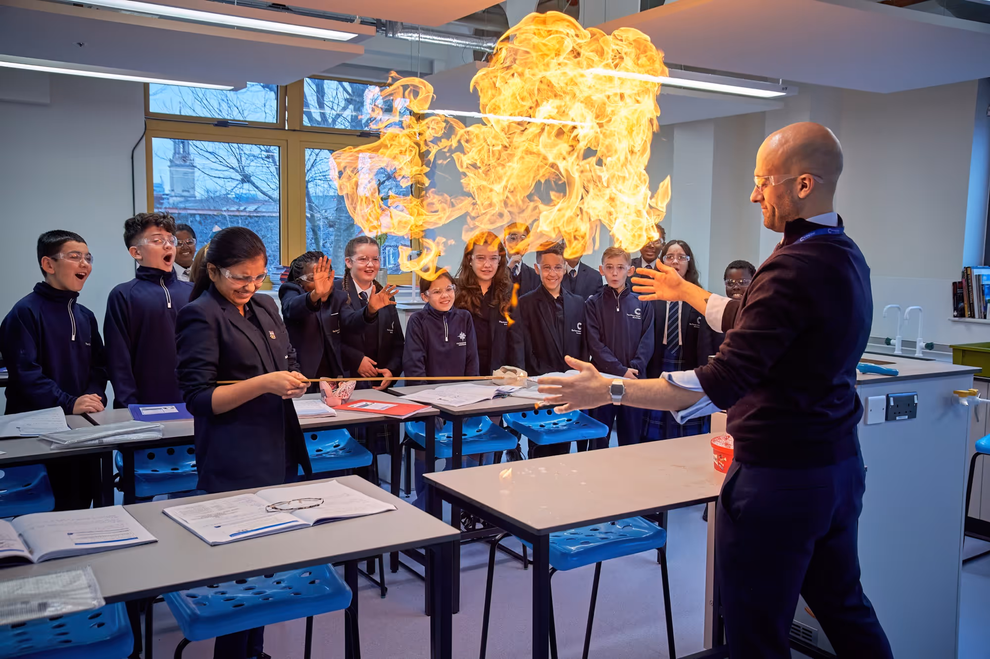 A science teacher performing a fire experiment in a classroom as a group of students watching attentively react with amazement while the flame bursts from a container held by the teacher.