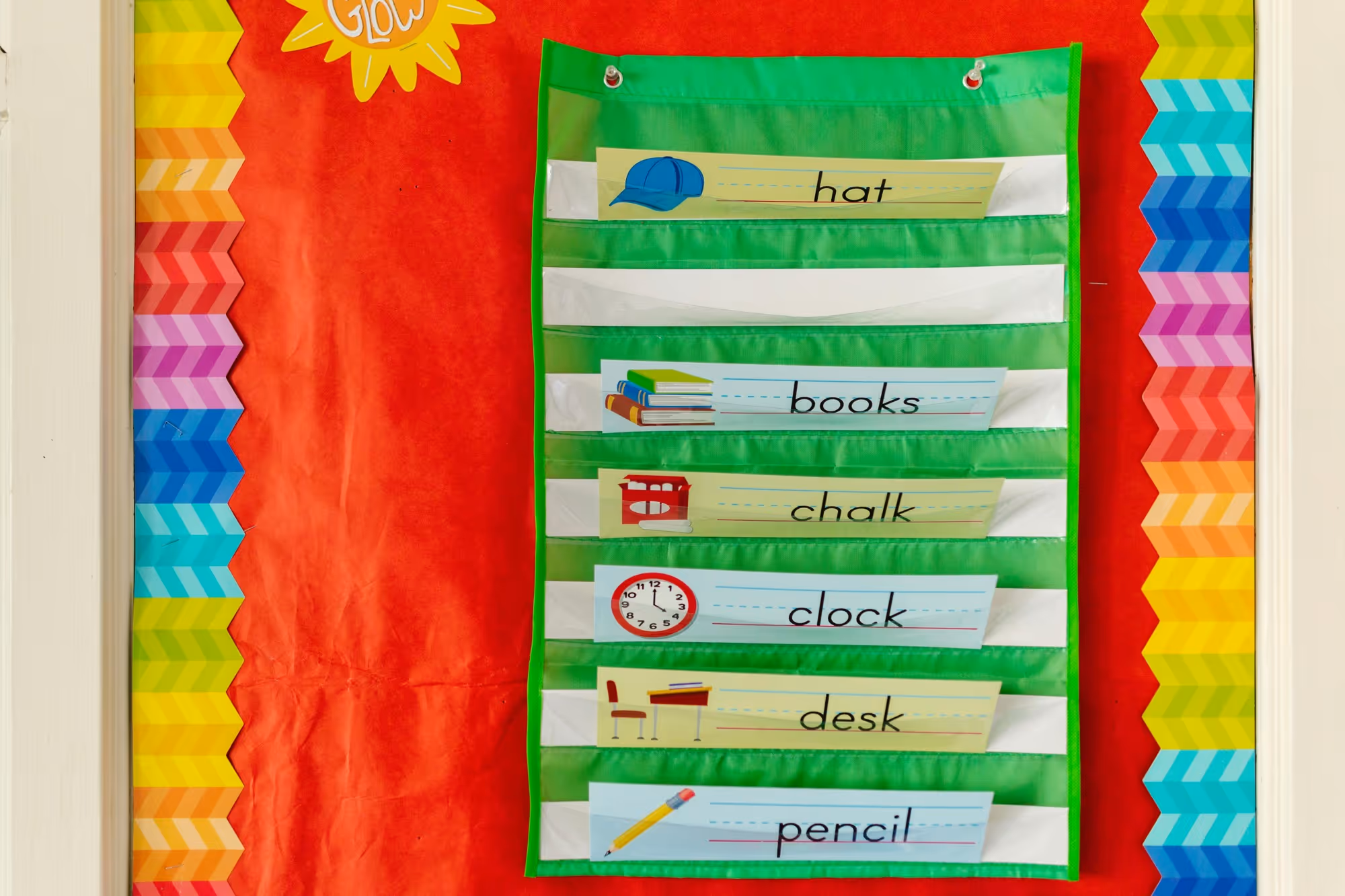 Classroom wall pocket chart with green and white striped pockets hanging on a red bulletin board with colorful zigzag border, showing labels and images for hat, books, chalk, clock, desk, and pencil.