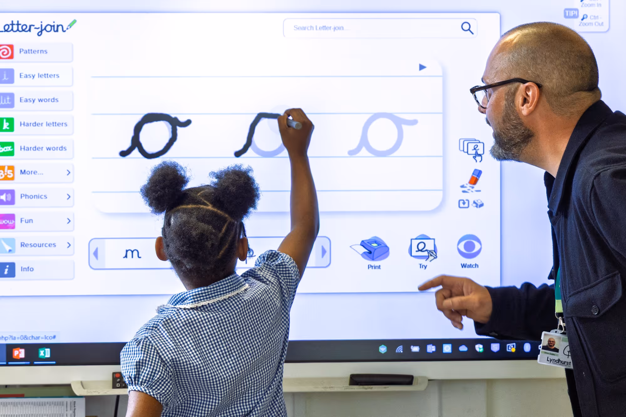 A young girl with puffs in her hair writes on a large interactive whiteboard while a man with glasses instructs her, pointing at the writing on the screen that shows a handwriting learning program with letters and patterns.