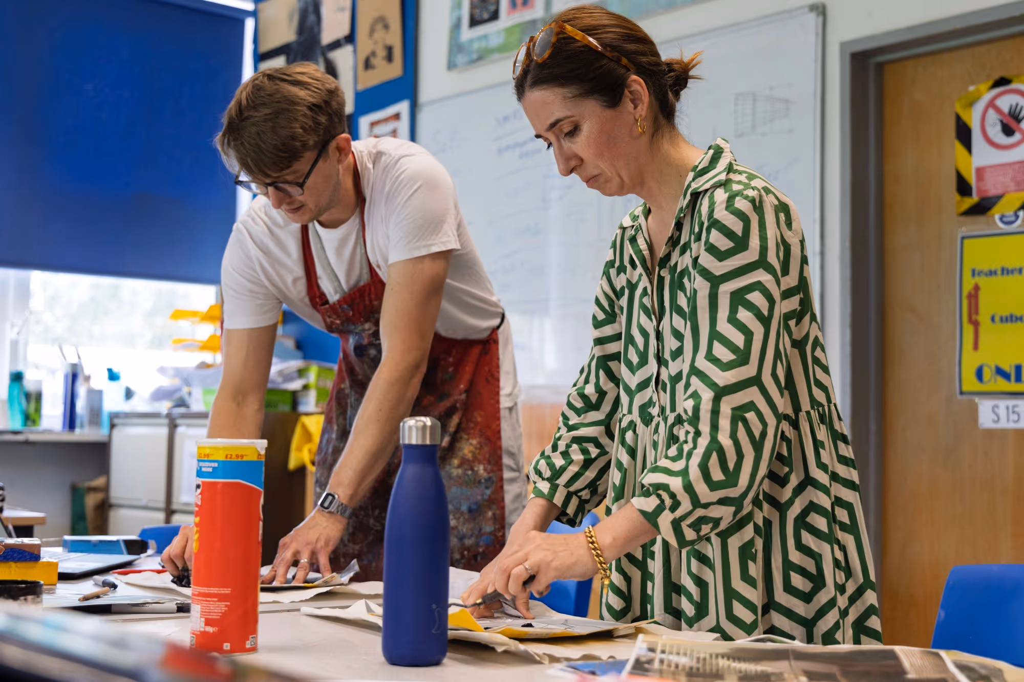 Two people working at a table covered with papers and documents in a brightly lit room, one wearing a green patterned dress and the other a white shirt and apron.