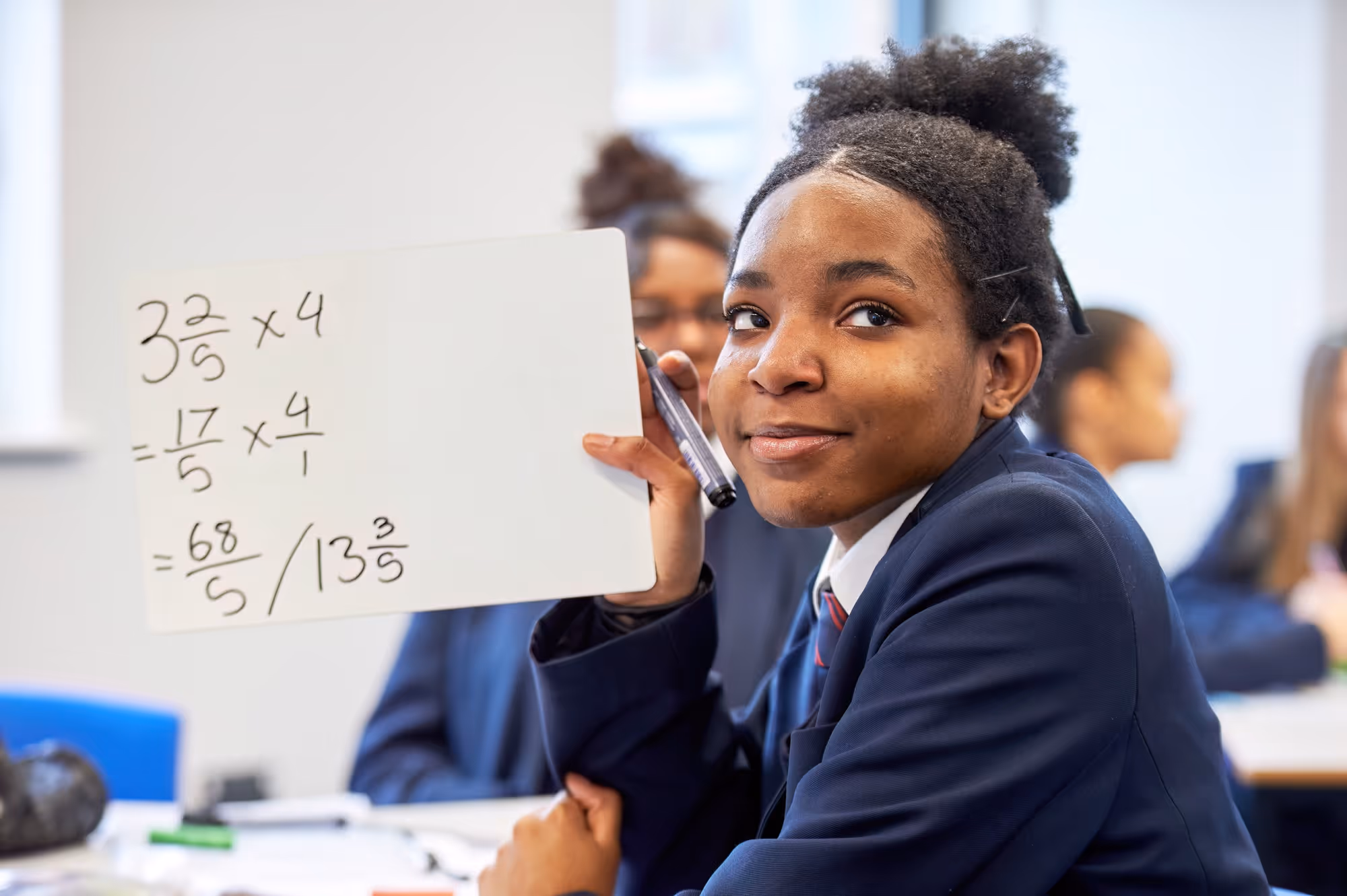 Student in school uniform holding up a whiteboard with a math problem involving multiplying mixed fractions written on it.