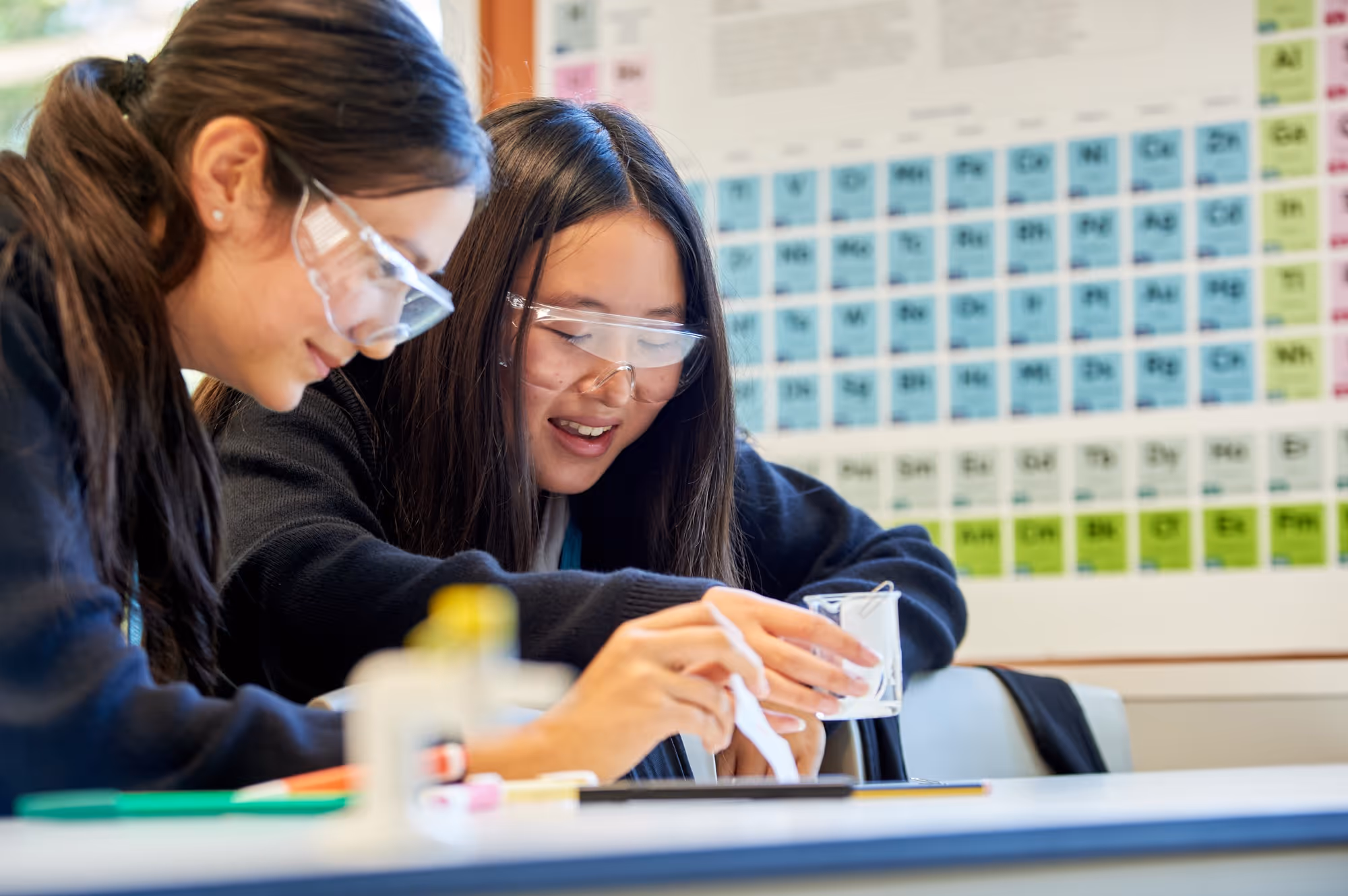 Two female students wearing safety goggles conducting a chemistry experiment in a classroom with a periodic table on the wall behind them.