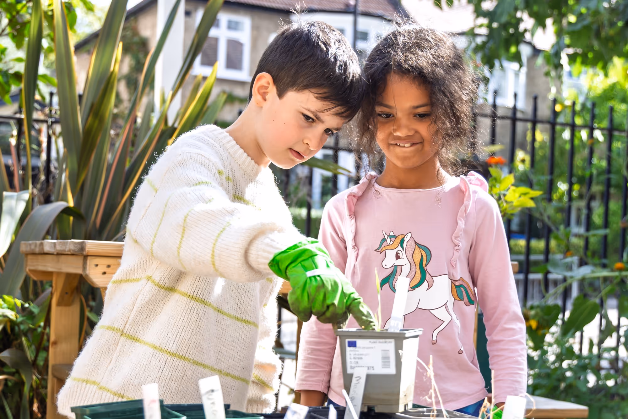 Two children gardening outdoors, one wearing a white sweater and green gloves planting in a pot, the other smiling and watching wearing a pink shirt with a unicorn print.