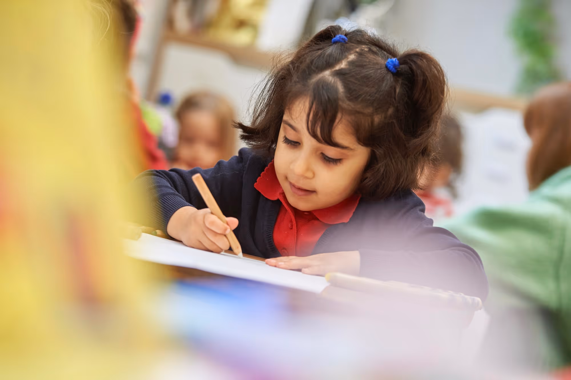 Young girl with dark hair in pigtails wearing a red shirt and dark sweater concentrating as she writes or draws on paper in a classroom setting with blurred children in the background.