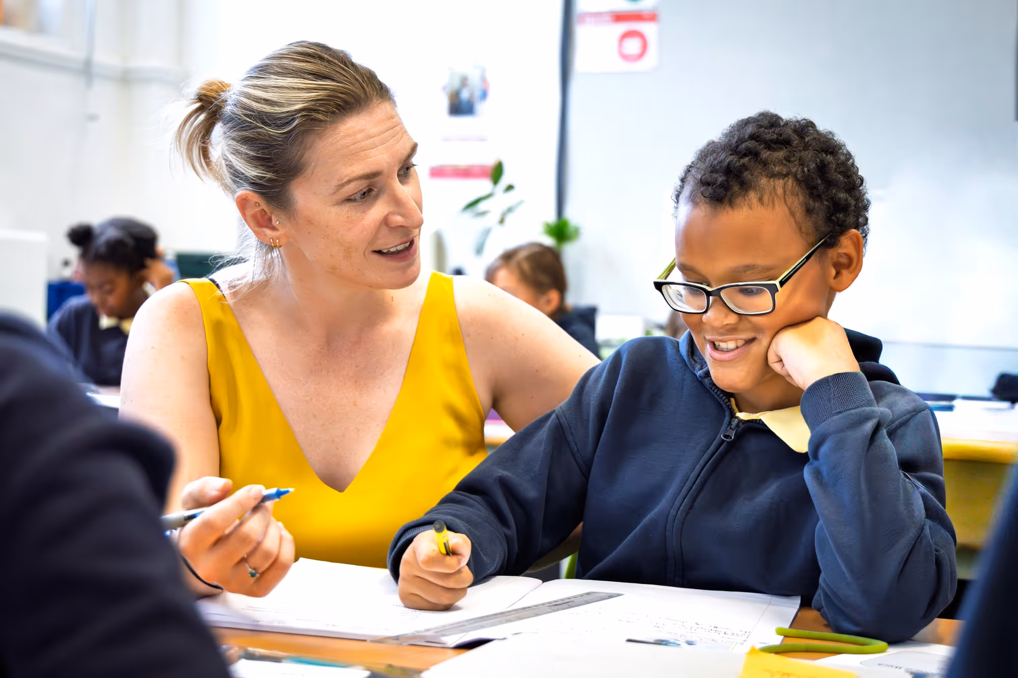 A female teacher in a yellow sleeveless top is helping a smiling young boy with glasses, seated at a desk with his writing materials in a classroom.