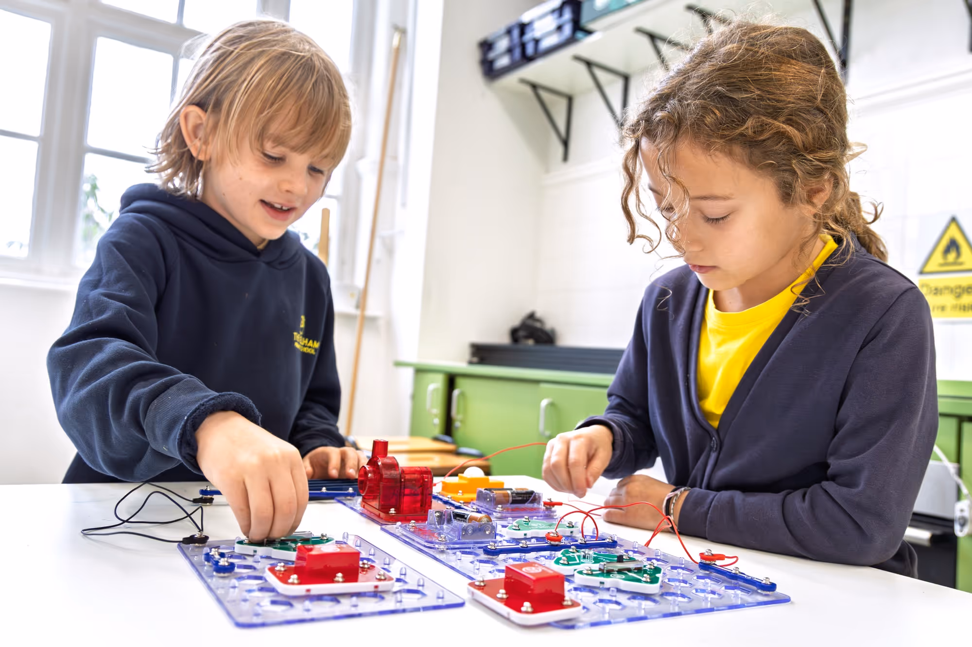 Two young children in a classroom setting engaged in assembling circuit boards with electronic components on a white table, with shelves and cabinets in the background.