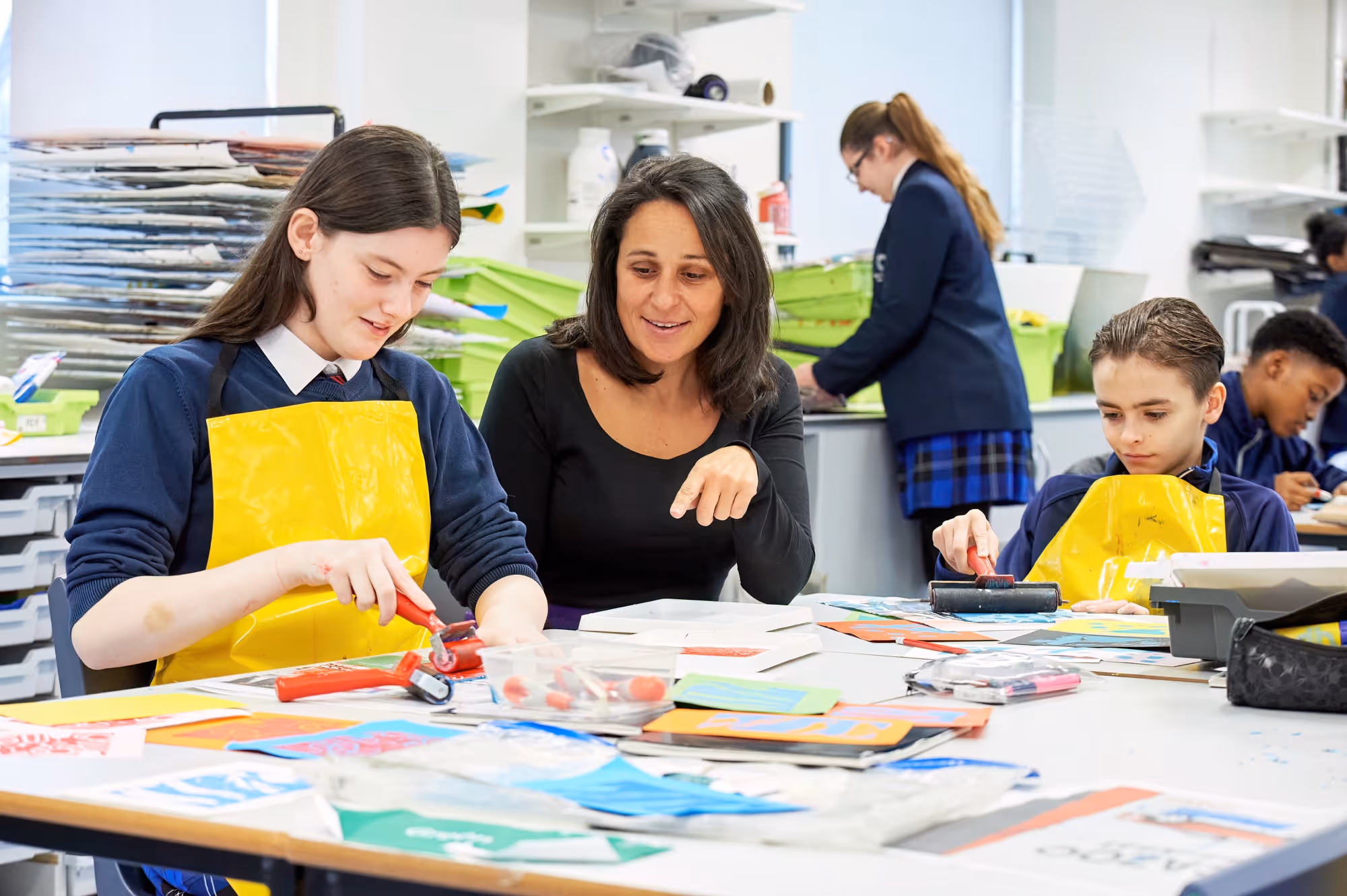 A teacher guides two children painting on paper in a bright classroom with art supplies and other students working in the background.