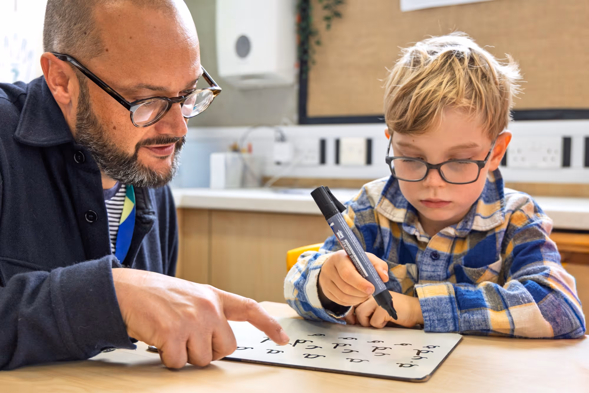 Teacher with glasses pointing to letters on a whiteboard as a young boy with glasses and a plaid shirt practices writing with a marker in a classroom.