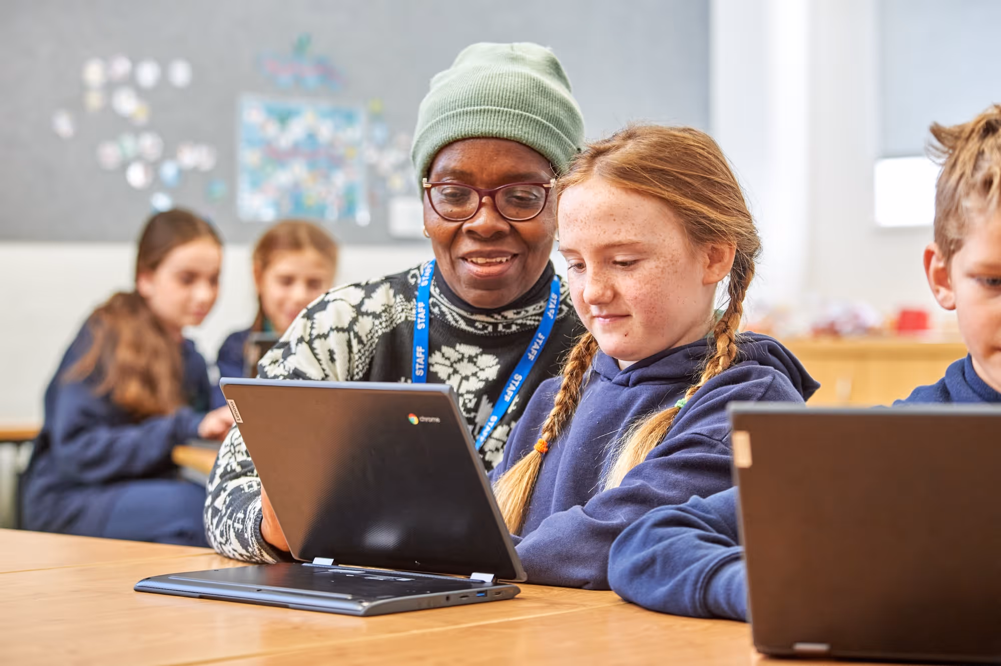 Teacher wearing glasses and a green beanie helps a young girl with braided hair who is using a Chromebook laptop in a classroom setting, with other students also engaged with laptops in the background.