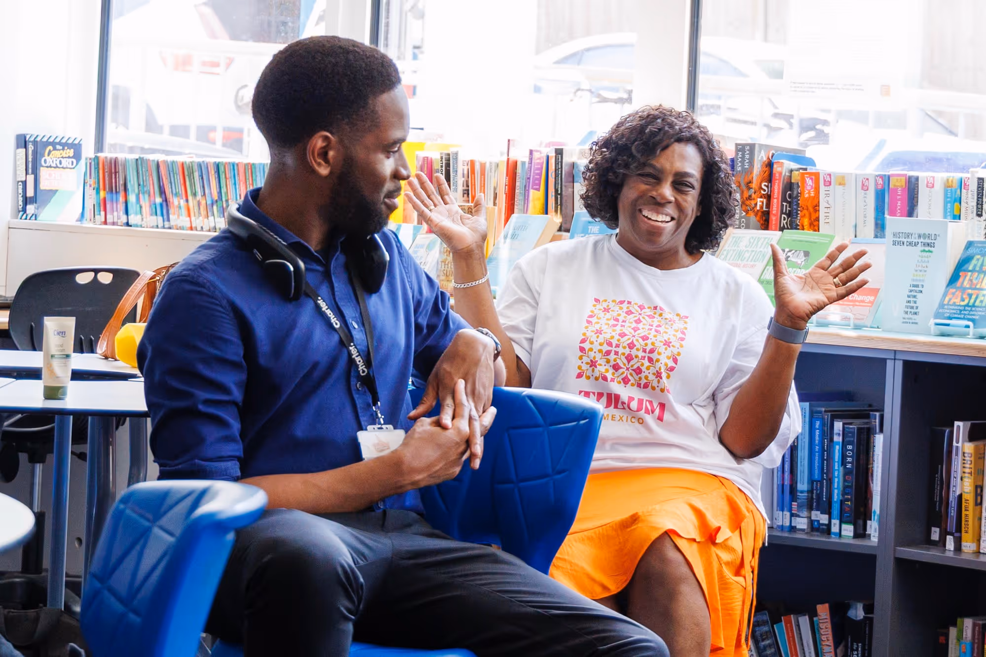 Smiling woman in a white T-shirt and orange skirt gestures with raised hands while talking to a man in a blue shirt with headphones around his neck in a bright room with bookshelves behind them.