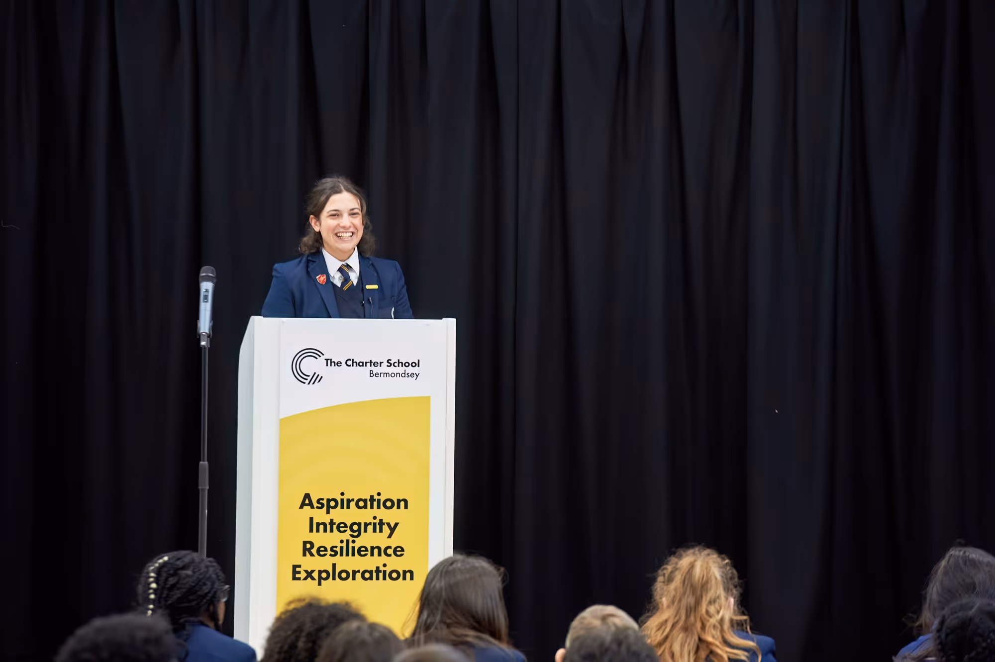 A female student in school uniform smiling while standing behind a podium with a microphone, addressing an audience against a black curtain background; the podium displays The Charter School Bermondsey logo and the words Aspiration Integrity Resilience Exploration.
