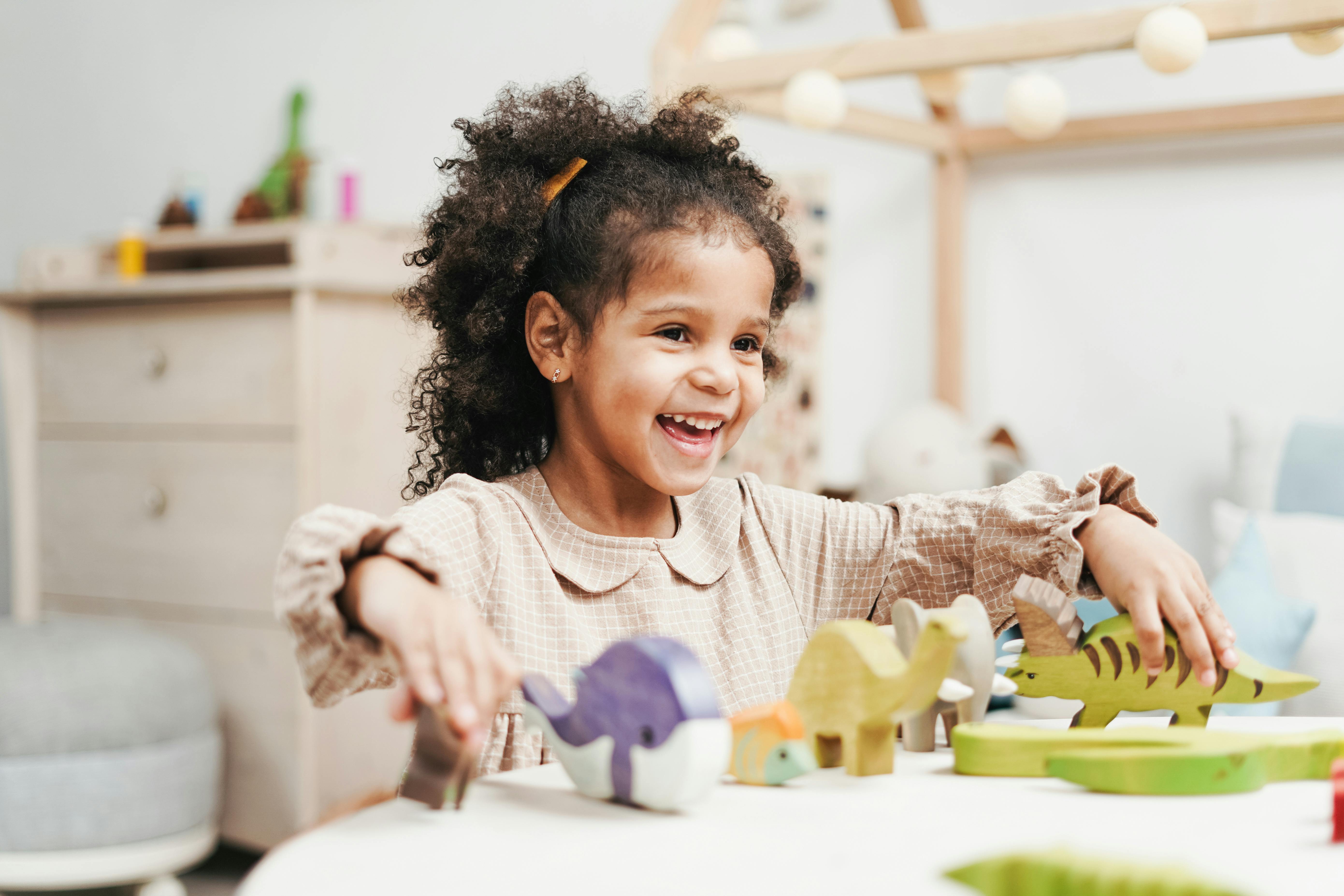 Smiling young girl with curly hair playing with colorful wooden animal toys on a white table in a bright room.