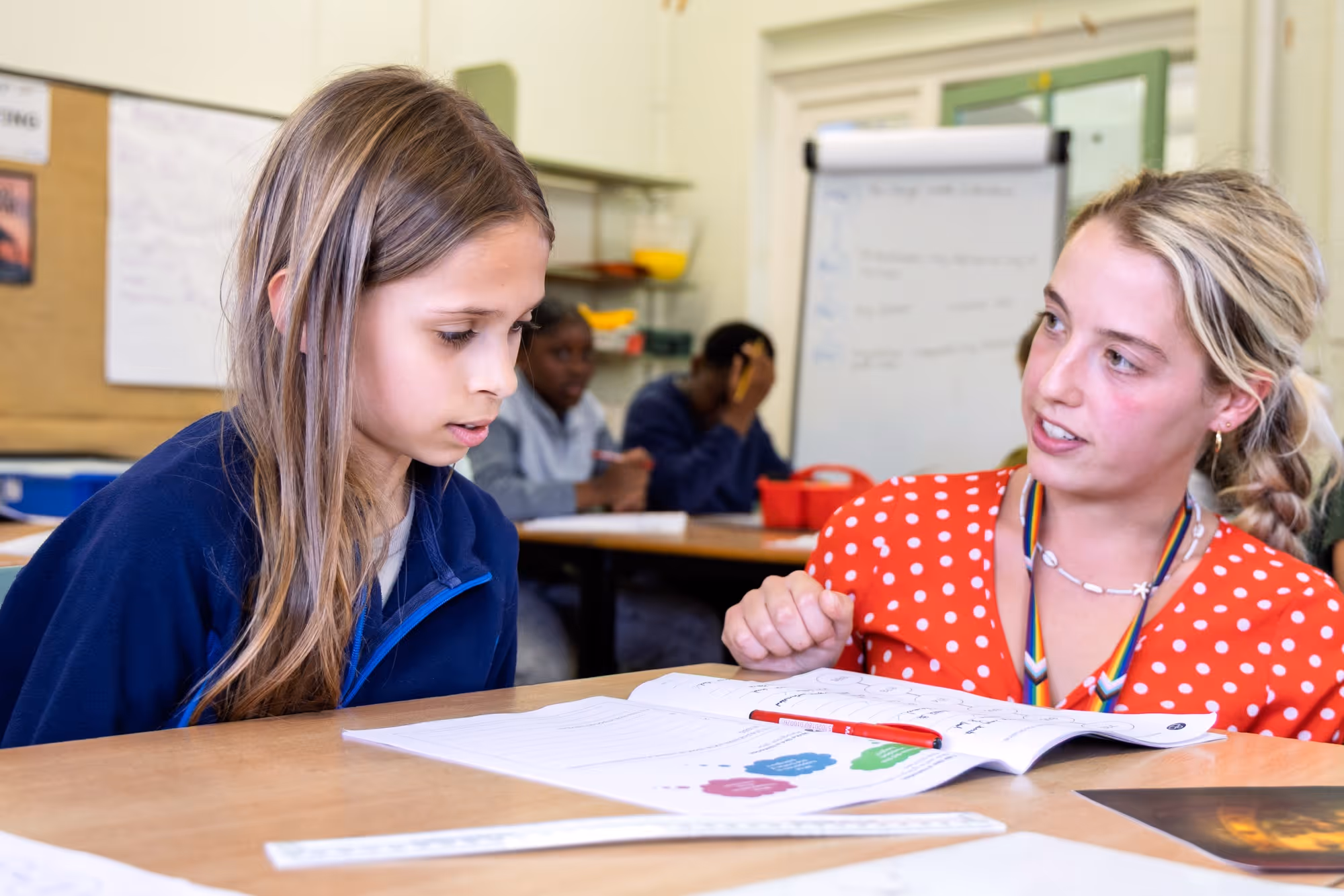 Young girl with long hair looking at an open workbook while a woman in a red polka-dot shirt explains something beside her in a classroom setting.