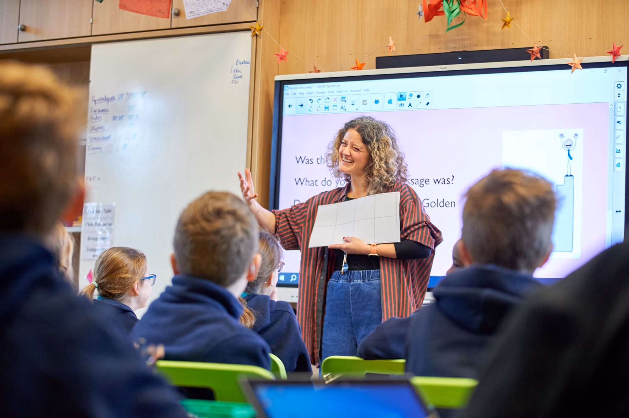 A female teacher with curly hair smiles and gestures while holding a paper grid in front of a classroom of attentive students, with a large screen behind her displaying a lesson with text and a cartoon figure on a podium.