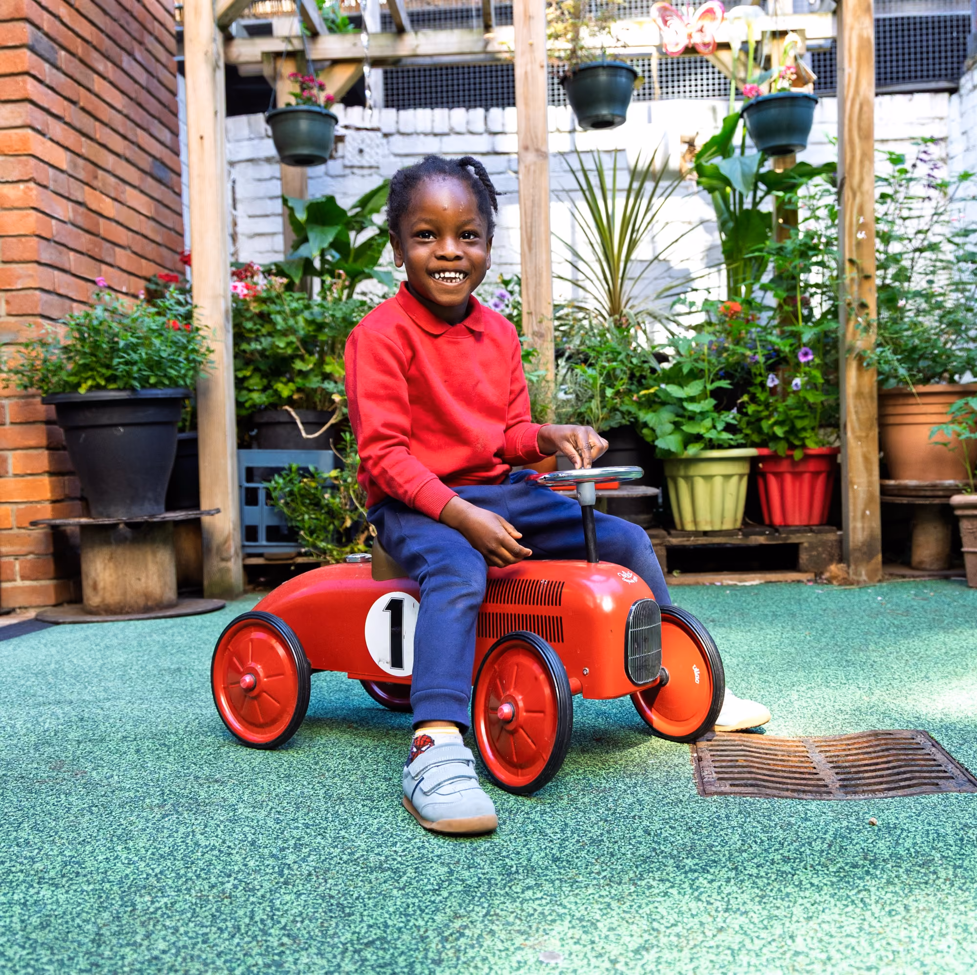 Smiling young child in a red sweater and blue pants riding a red toy car with the number 1 on it in a garden area with potted plants and a brick wall.