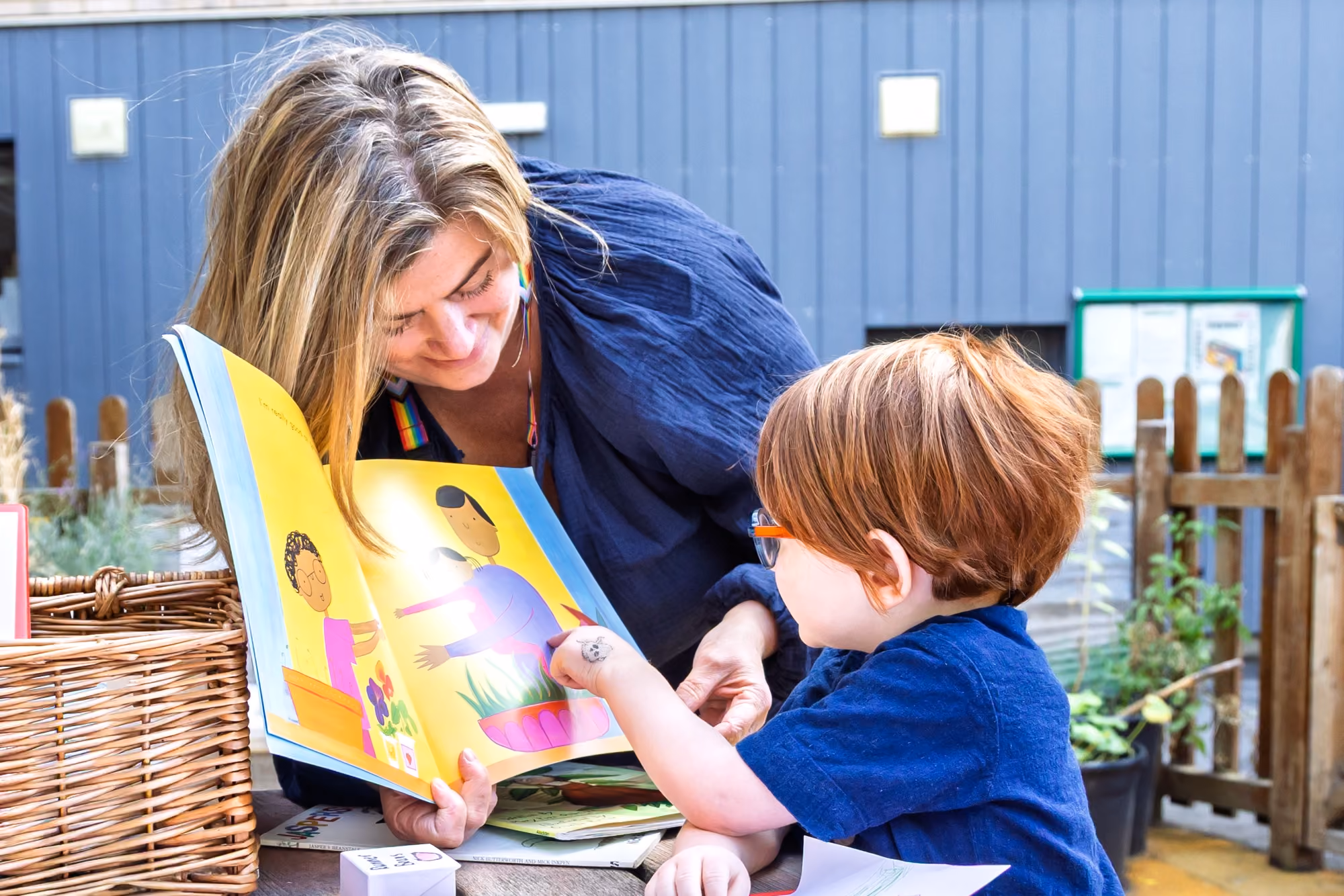 Woman with long blonde hair and a rainbow strap on her shirt reading a colorful children's book with illustrations to a young child with short red hair and glasses who is pointing at the book outdoors near a wooden fence and wicker basket.
