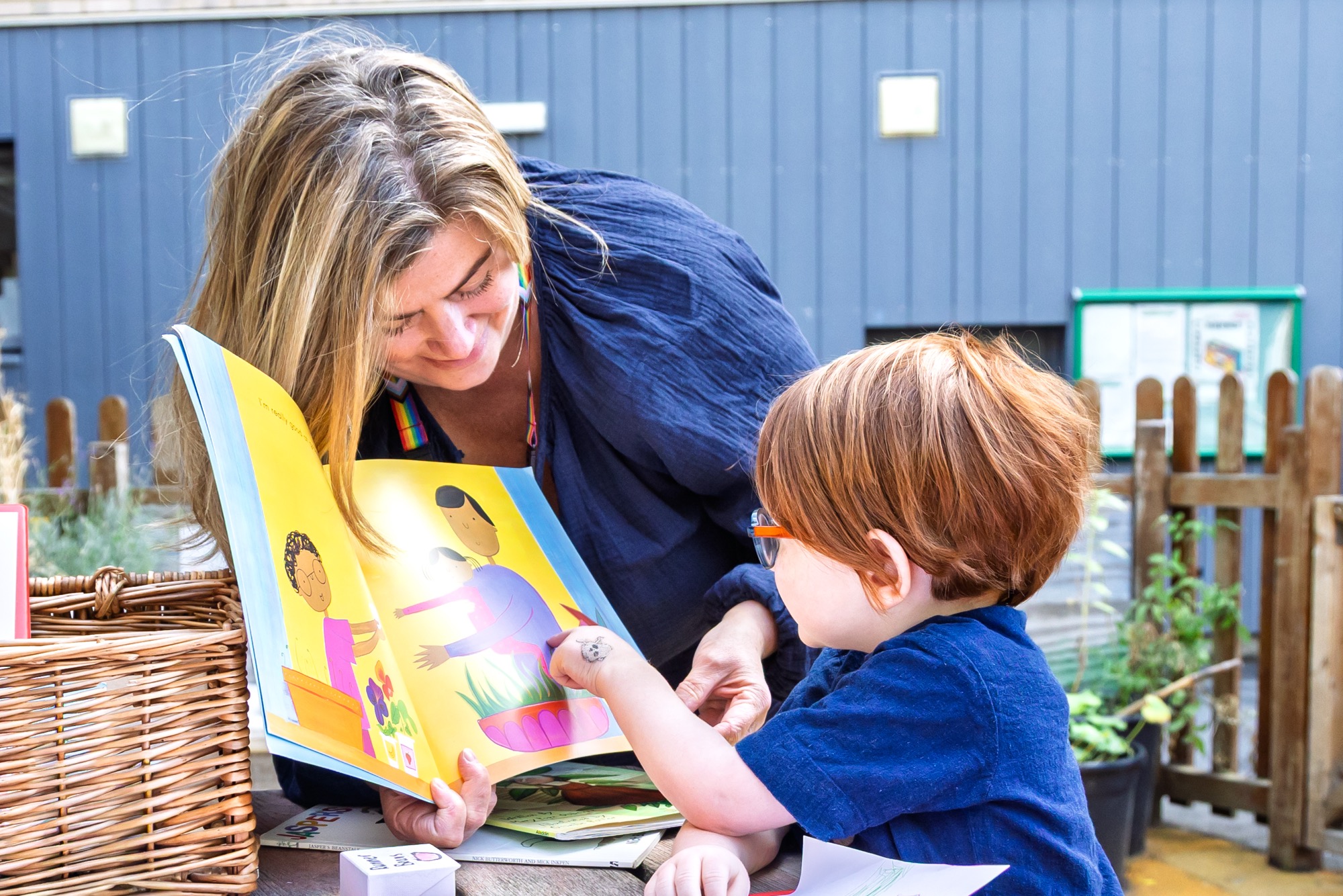 Woman with long blonde hair and a rainbow strap on her shirt reading a colorful children's book with illustrations to a young child with short red hair and glasses who is pointing at the book outdoors near a wooden fence and wicker basket.