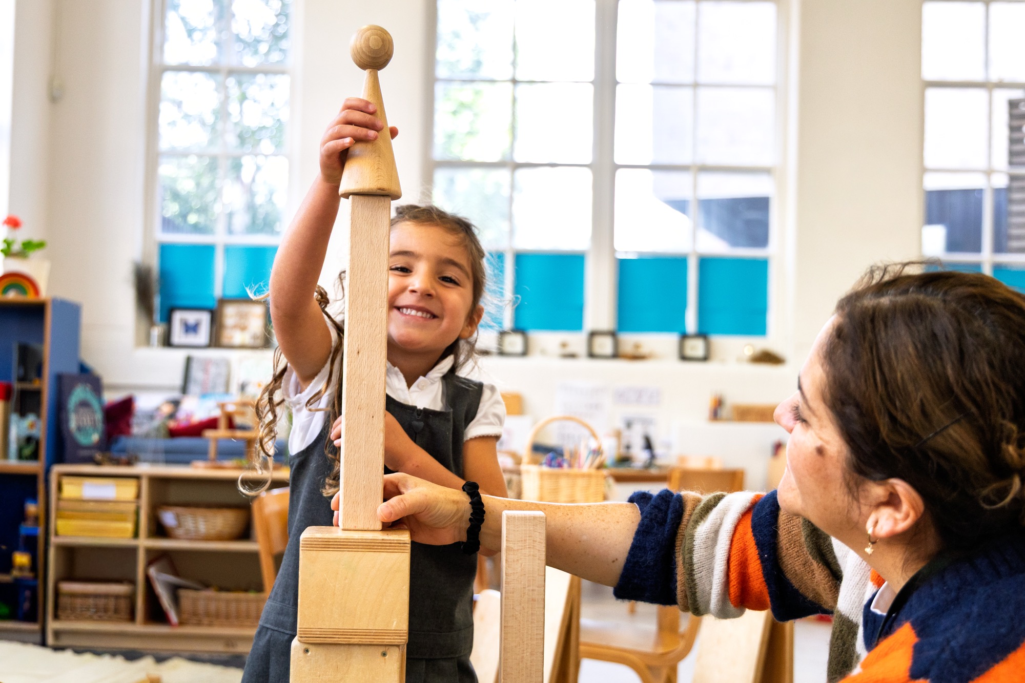 A young girl smiling while holding up a tall wooden block tower with the help of an adult woman in a brightly lit classroom.
