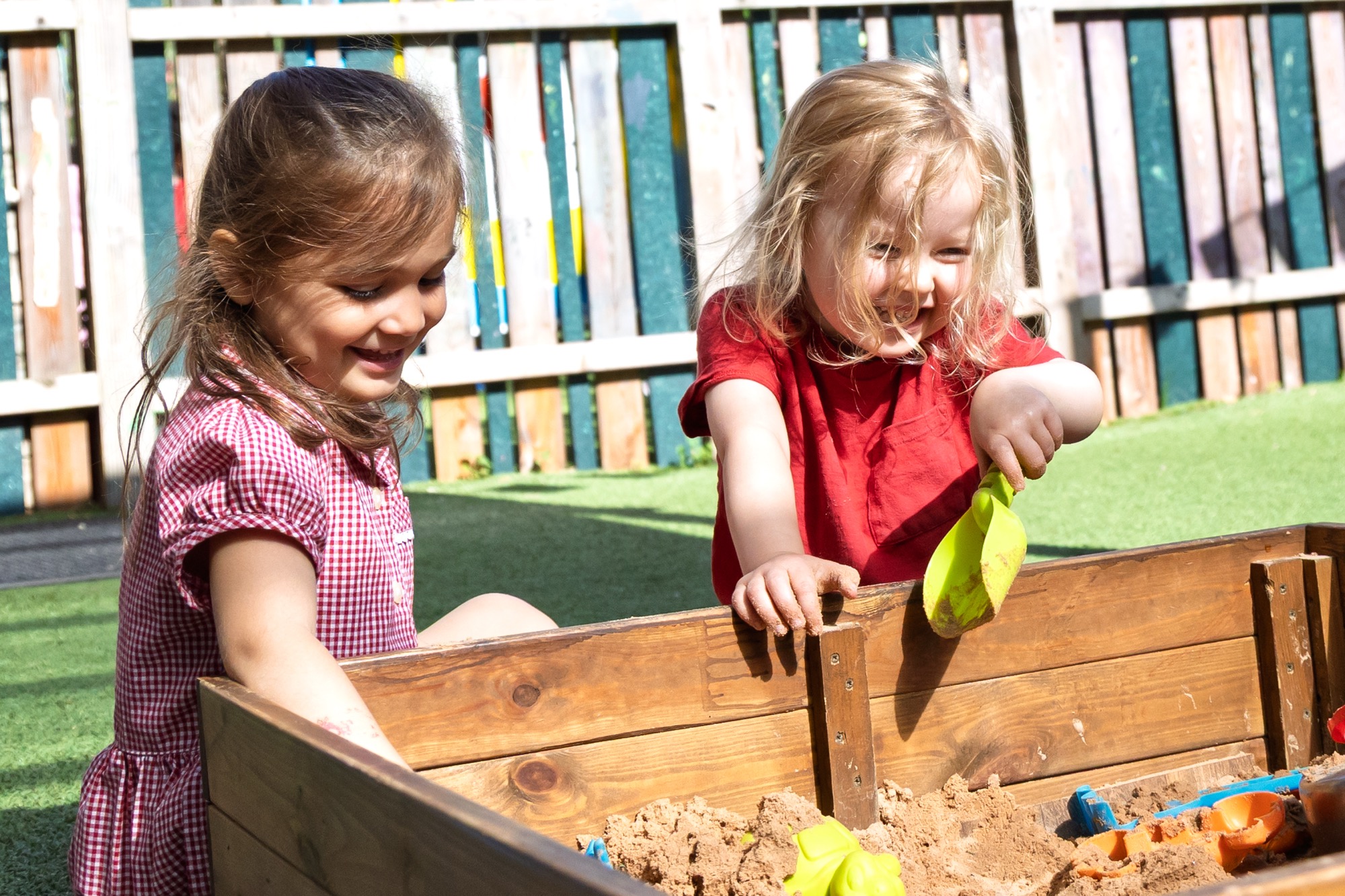 Two young girls playing and smiling happily in a wooden sandbox filled with sand and toys, outdoors on green artificial grass with a colorful wooden fence in the background.