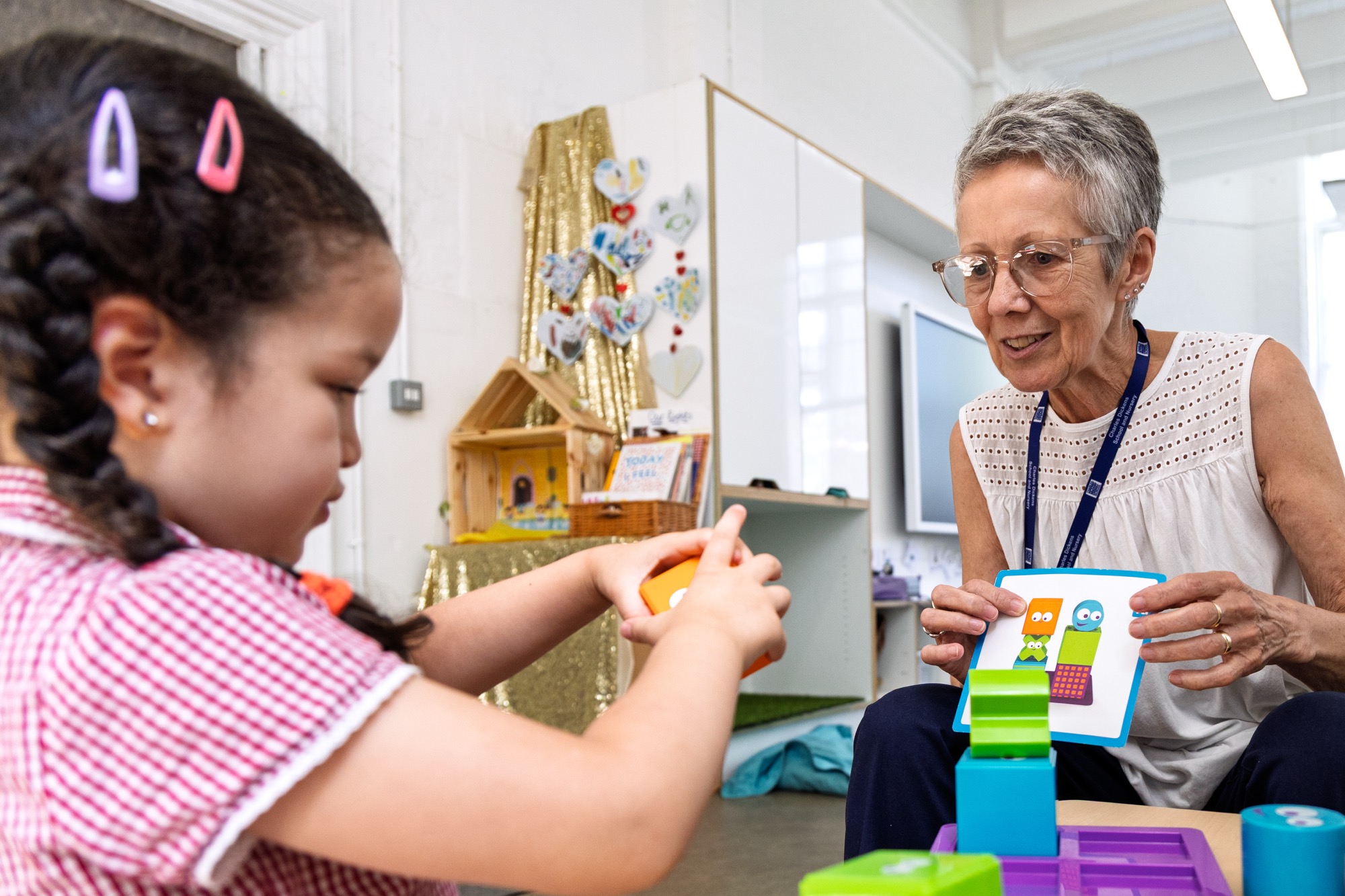 Young girl with braided hair and hair clips playing with colorful blocks while an older woman with short gray hair and glasses holds a card with cartoon block characters, sitting together in a bright classroom or nursery setting.