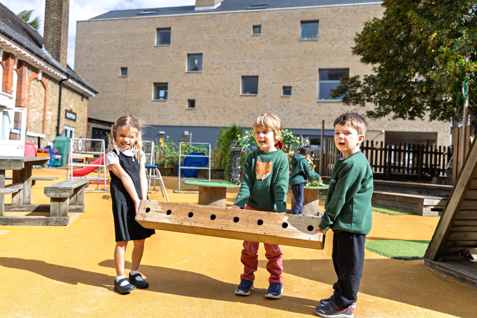 Three children standing outdoors on a playground, holding a large wooden plank with holes, with a building and playground equipment in the background.
