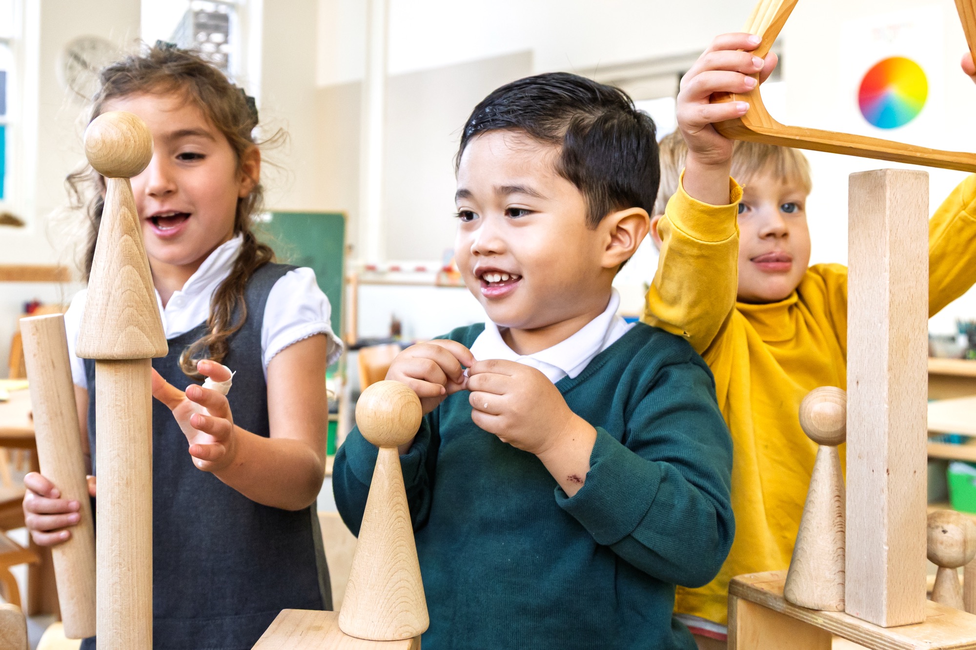 Three young children in a classroom playing with large wooden shapes, building a structure together and looking engaged and happy.