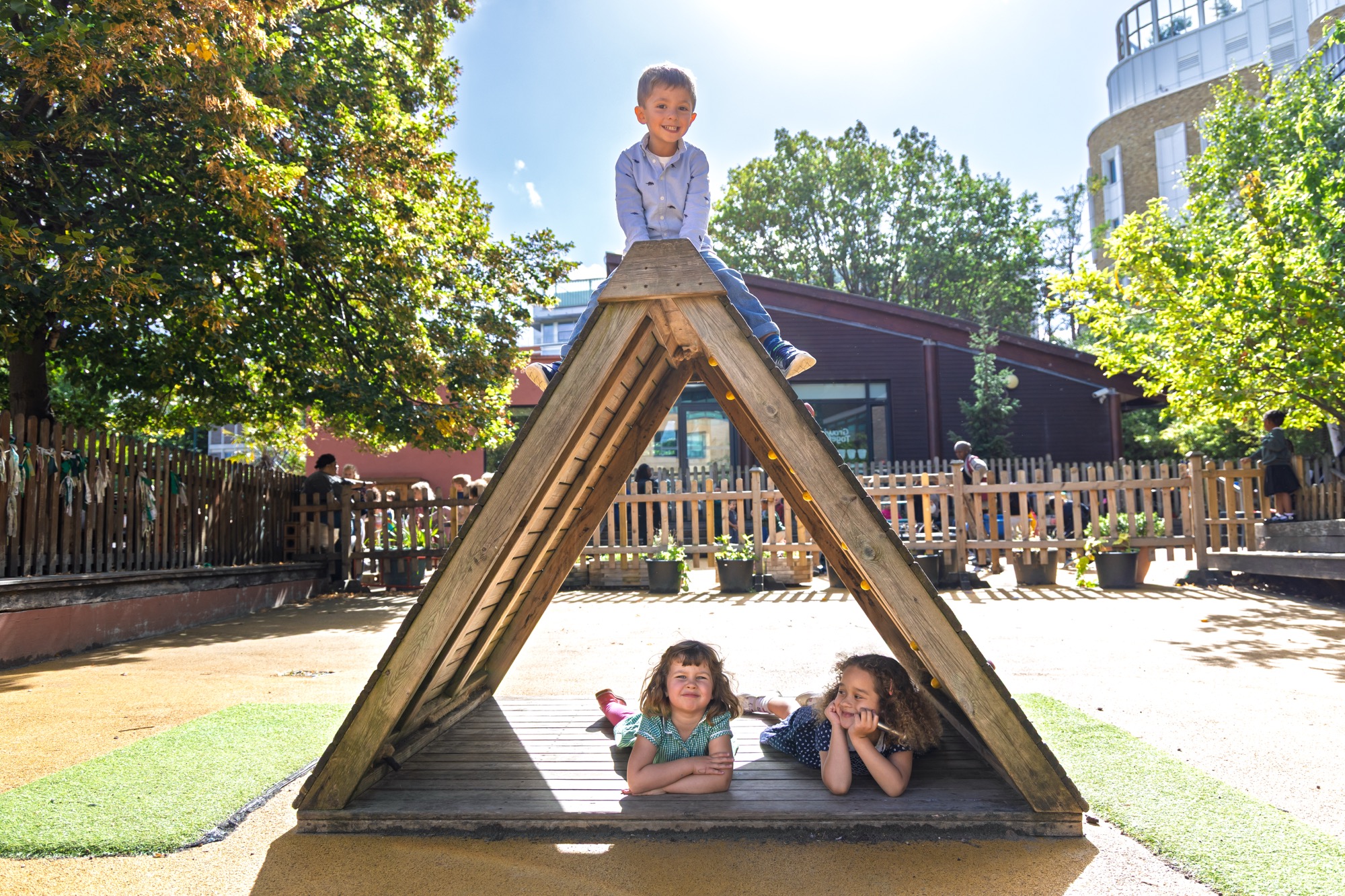 Three children playing on a wooden triangular play structure in a sunny outdoor playground with trees and a fenced area in the background; one boy sits atop the structure smiling, two girls lie underneath on the wooden platform.