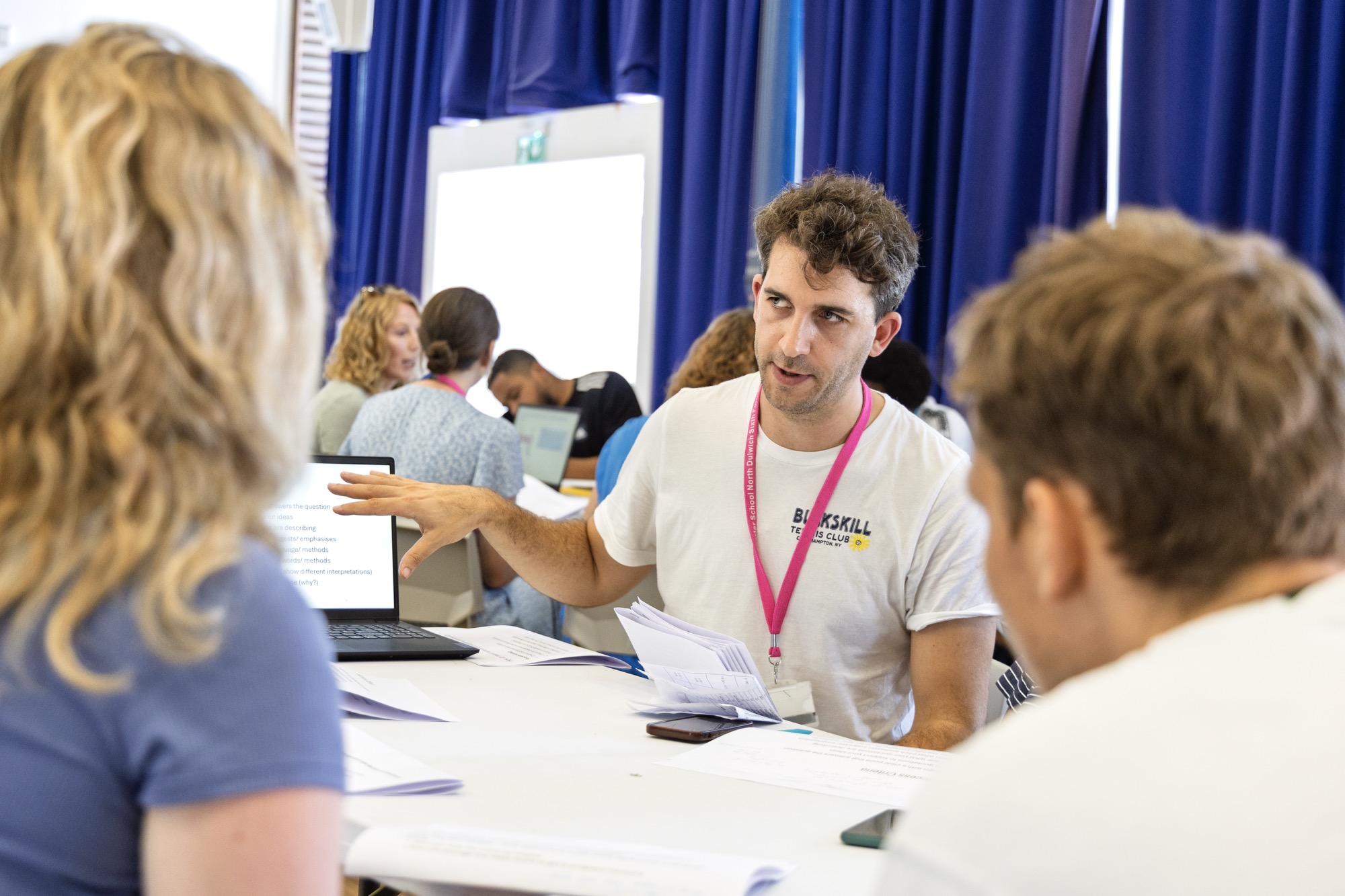 A man wearing a white T-shirt and pink lanyard is explaining something to two people sitting across a table, pointing at a laptop screen with text, in a room with blue curtains and other people working in the background.