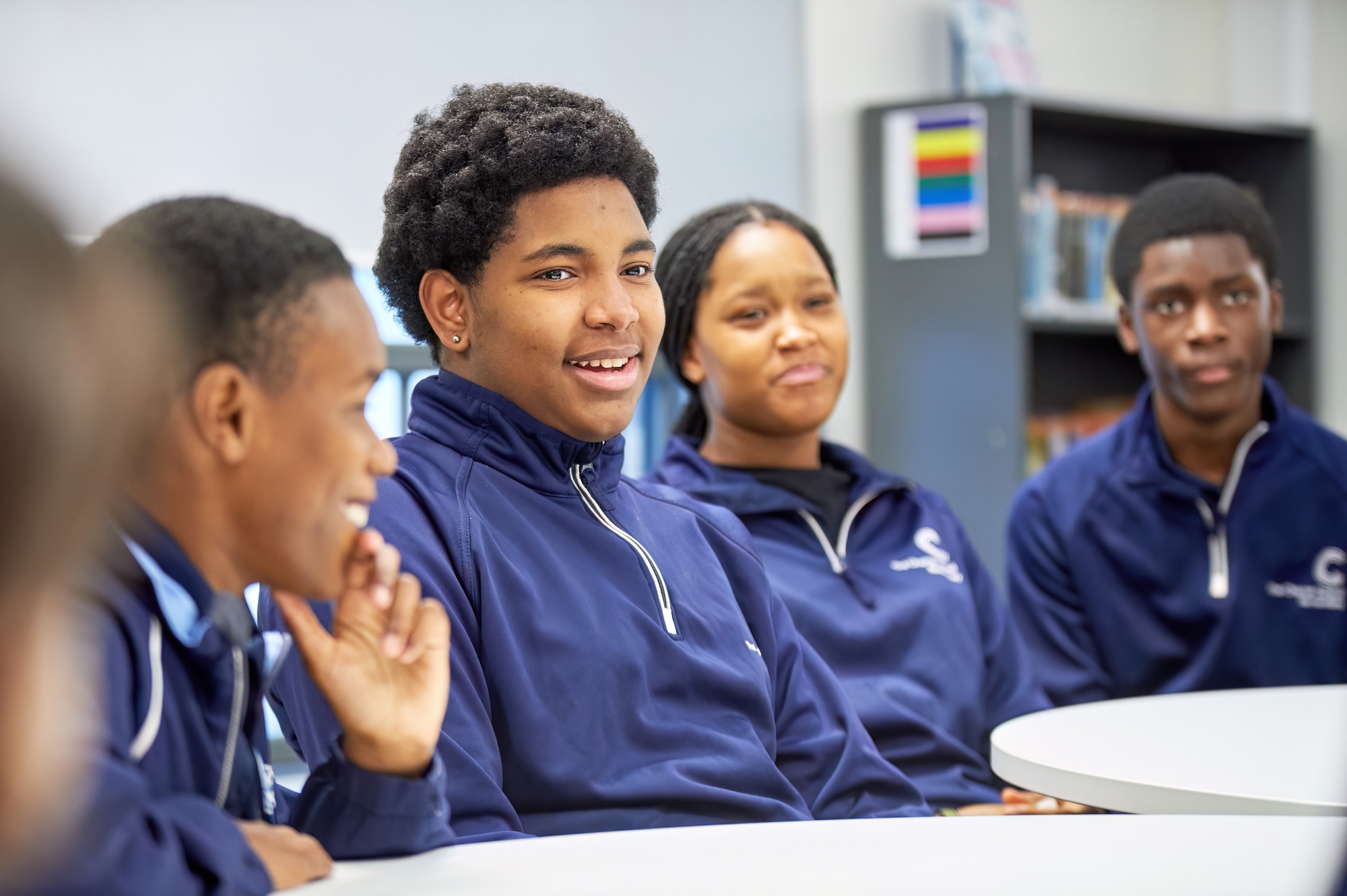 Four teenagers in blue jackets sitting around a white table, engaged in conversation in an indoor setting with shelves and colorful items in the background.
