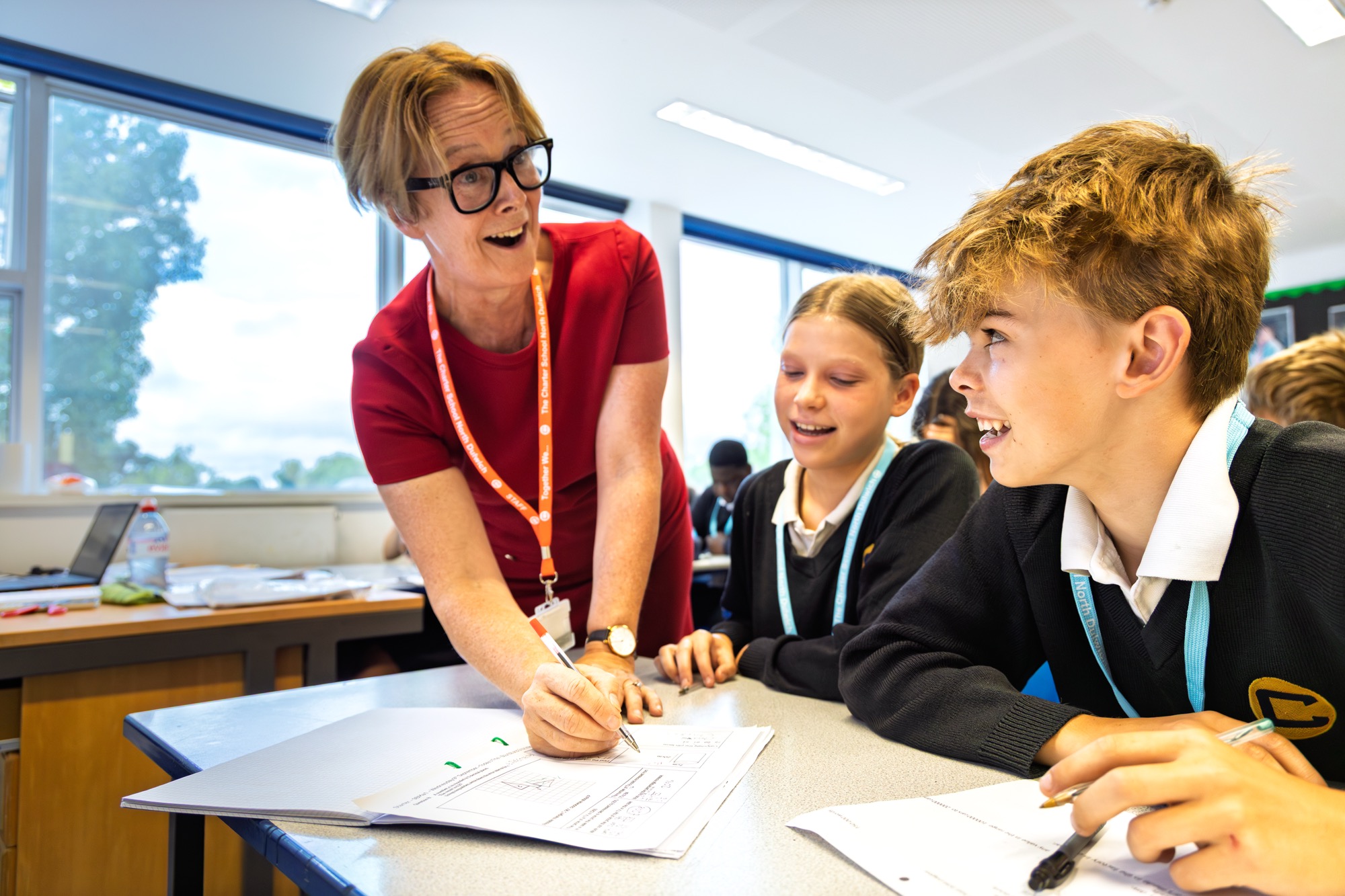 A teacher with glasses and a red dress is engaging with two smiling students in school uniforms at a desk, reviewing papers in a bright classroom with large windows.