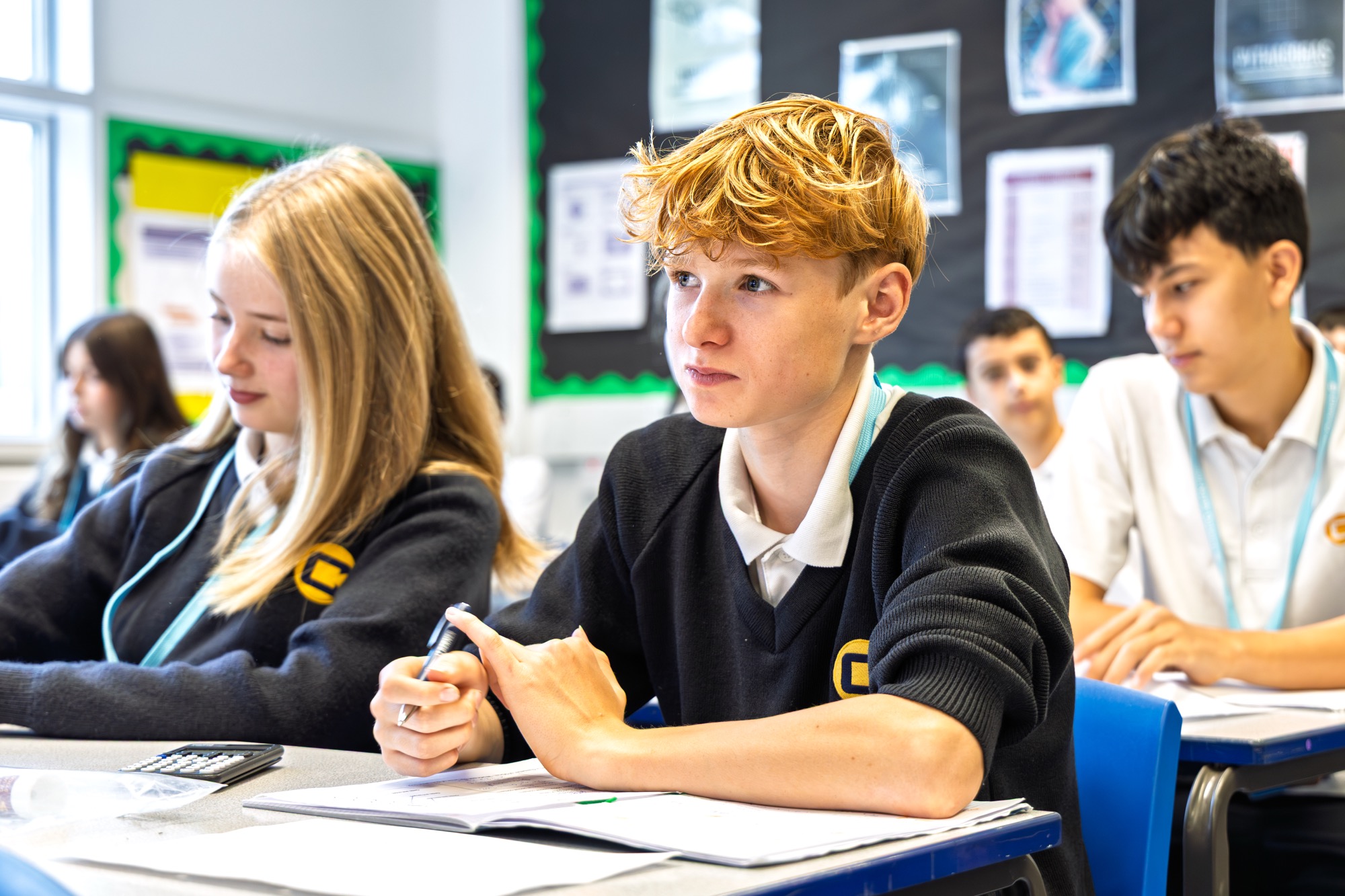 Classroom scene with focused teenage students sitting at desks, wearing school uniforms with black sweaters and white shirts, open notebooks, and a calculator visible on a desk.