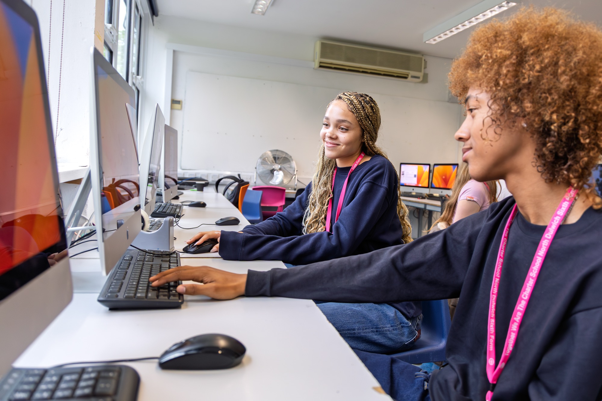 Two students sitting side by side at a long desk in a computer lab, working on desktop computers and wearing dark sweatshirts with pink lanyards, smiling and interacting casually.
