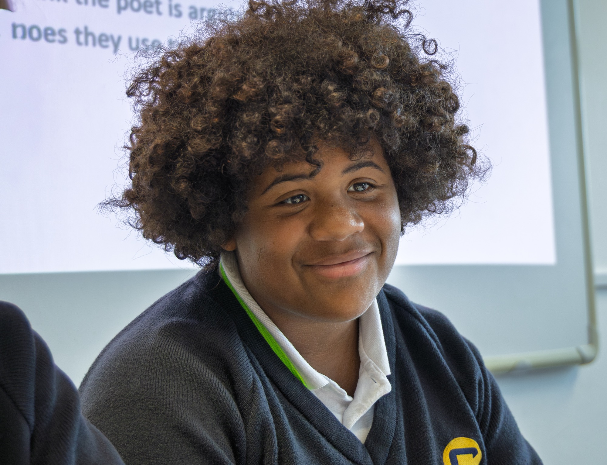 Smiling young person with curly hair wearing a dark sweater and white collared shirt in a classroom setting with a blurry whiteboard in the background.