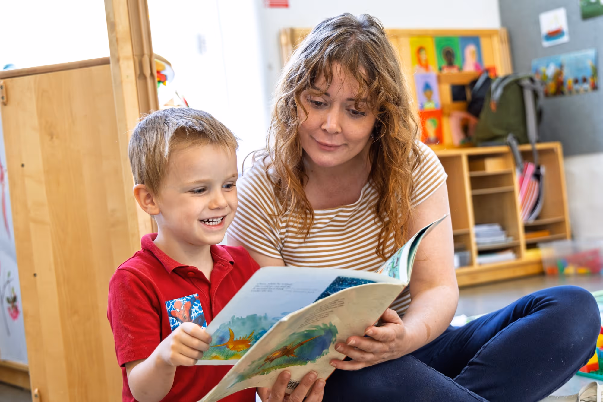 A woman with curly hair in a striped shirt sits with a smiling young boy in a red shirt, reading a colorful children's book together in a bright playroom with shelves and toys.