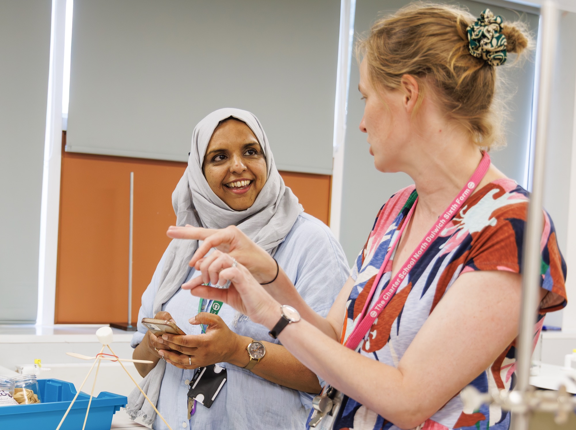 Two women engaged in conversation in a classroom setting; one is wearing a light gray hijab and holding a smartphone, while the other wears a colorful shirt with a pink lanyard that reads 'The Charter School North Dulwich Sixth Form,' gesturing with her hands.