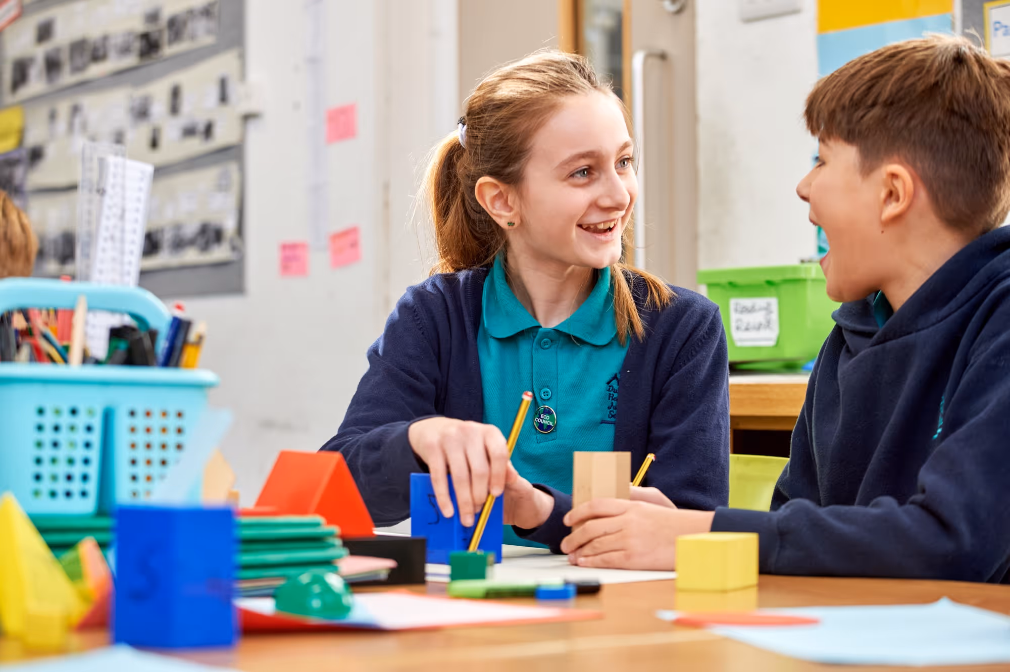Two children, a girl and a boy, sitting at a table in a classroom smiling and interacting while working with colorful geometric shapes and pencils.