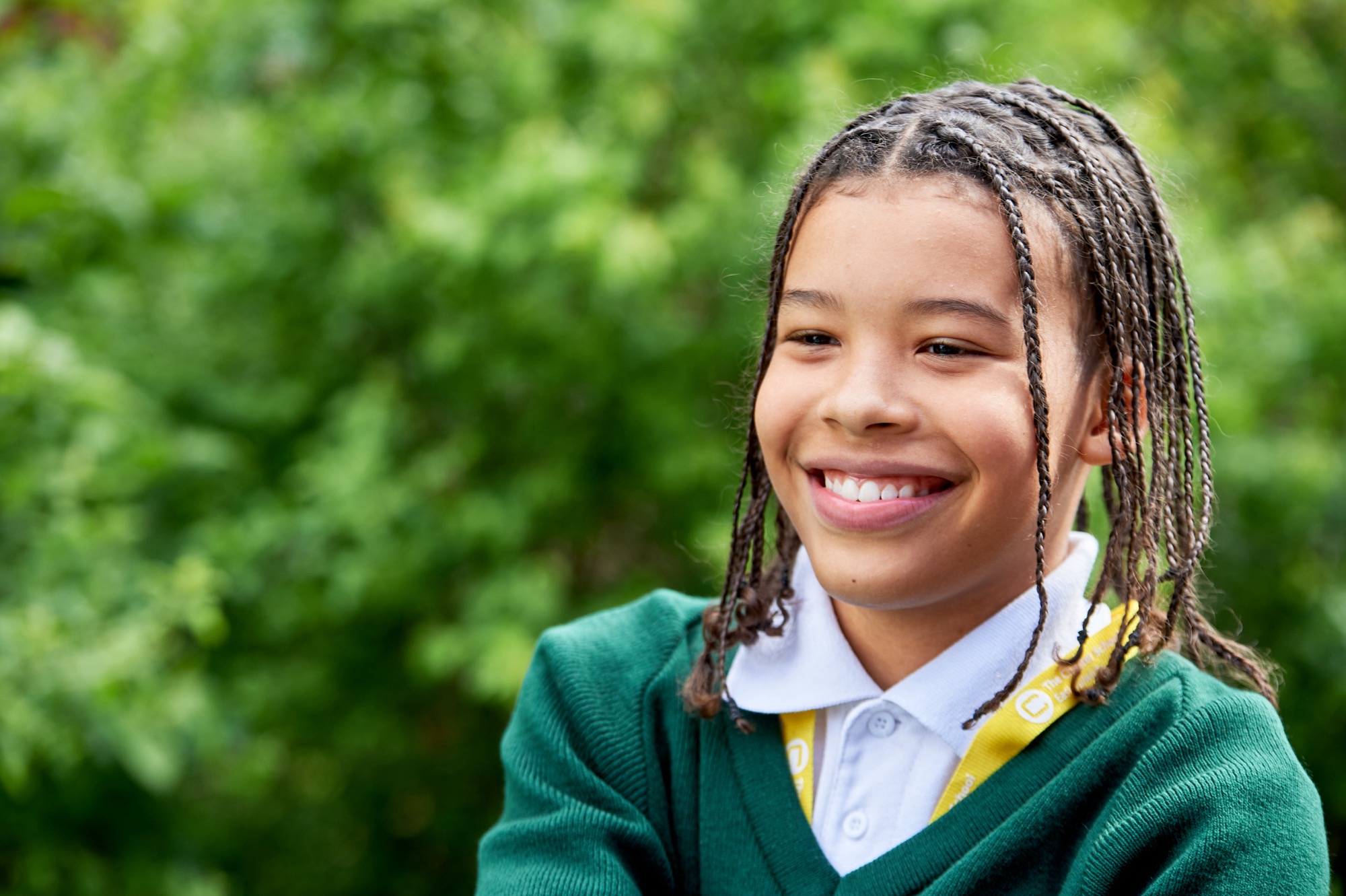Smiling child with braided hair wearing a green sweater and white collared shirt outdoors with green foliage in the background.