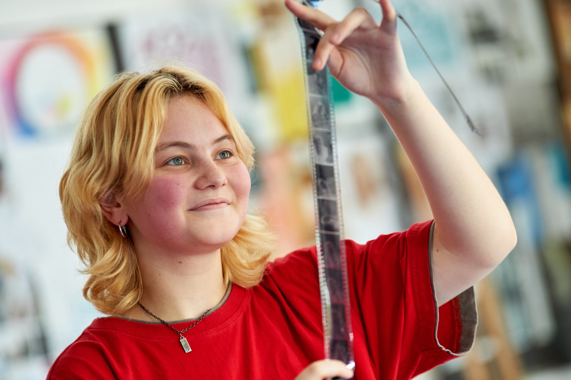 A young person with blonde hair and green eyes wearing a red shirt holds up and examines a strip of photographic film in a well-lit room with colorful blurred background.