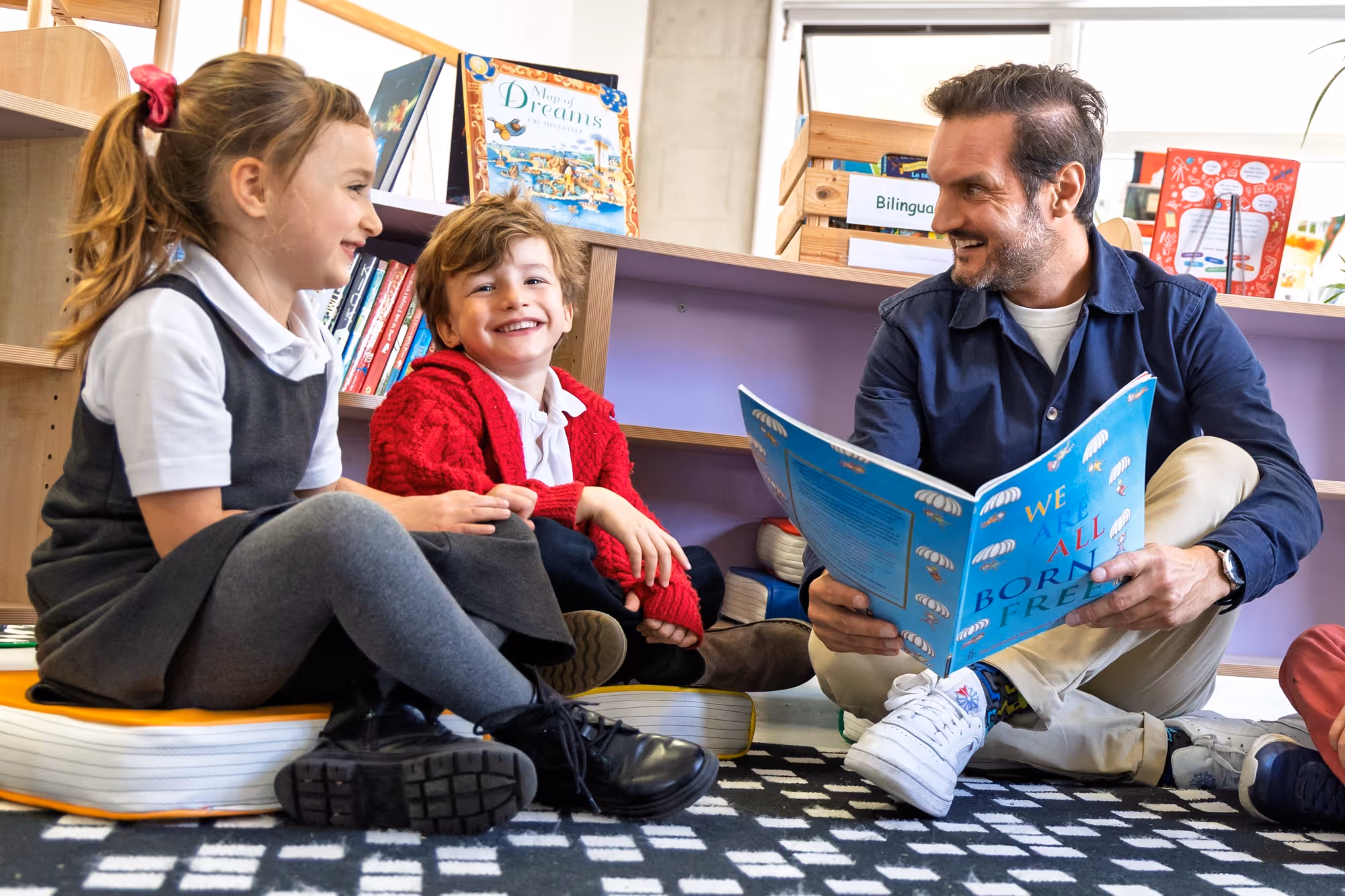 A man sitting on the floor reading a book titled 'We All Born Free' to two smiling children sitting on cushions in a cozy classroom area with bookshelves in the background.