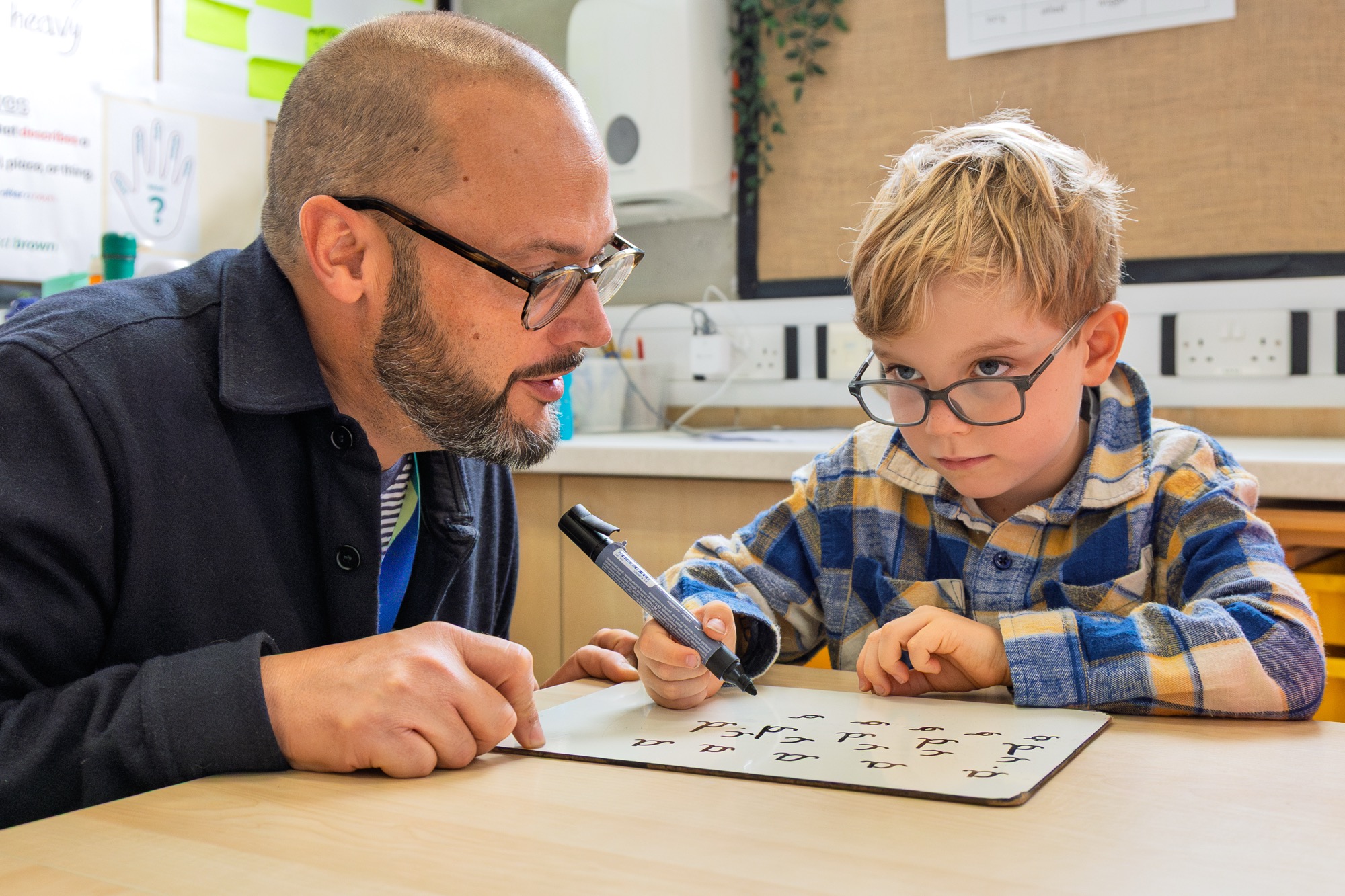 A young boy wearing glasses and a plaid shirt practices writing on a whiteboard while a man with glasses and a beard attentively watches and guides him in a classroom setting.