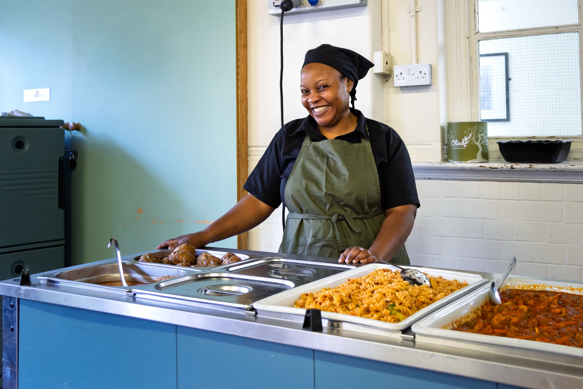 Smiling woman in a black shirt, green apron, and black headscarf serving food behind a buffet counter with trays of pasta, stew, and baked items.