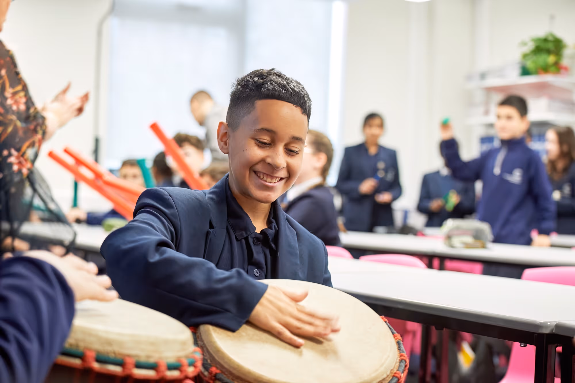 A smiling boy in a school uniform plays a hand drum in a classroom with other students, some holding colorful percussion instruments.