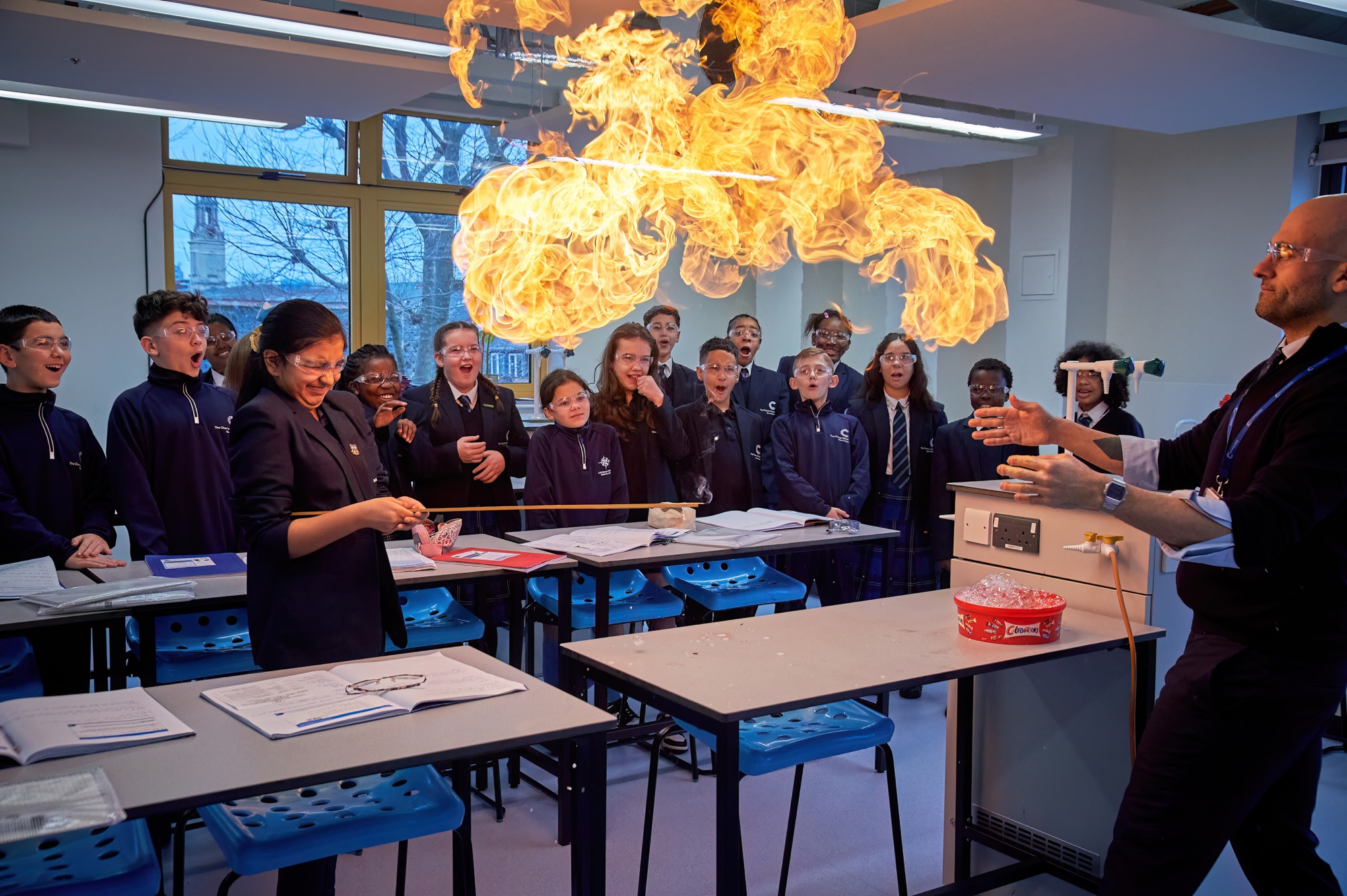 A science classroom where a teacher is demonstrating a fire experiment, creating a large flame above a table while a group of students watch in awe, all wearing safety goggles.