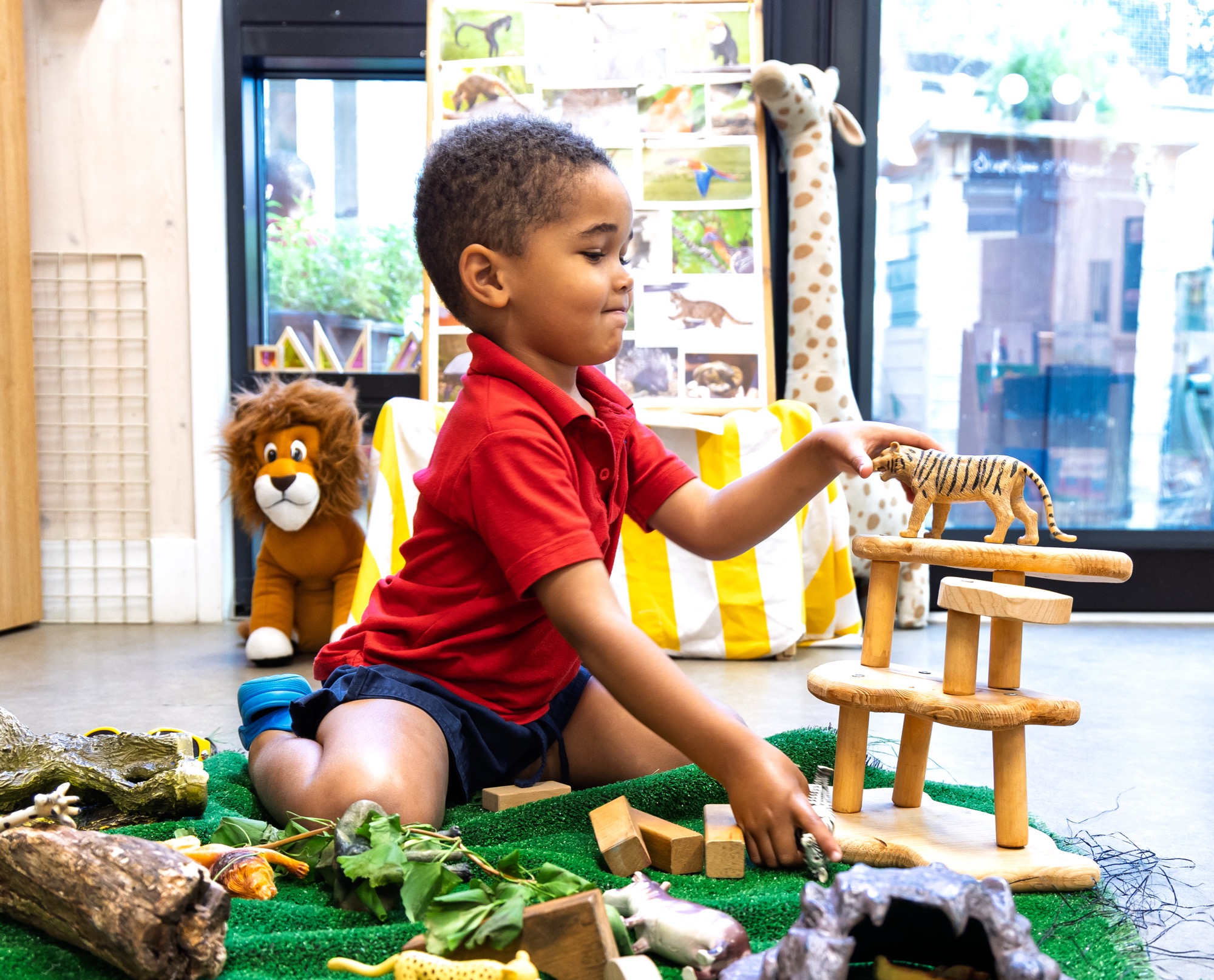 A young child wearing a red shirt and navy shorts sits on the floor playing indoors, stacking wooden blocks and placing a toy tiger on top, surrounded by other toy animals on green carpet, with a stuffed lion and a tall giraffe toy in the background near a window.