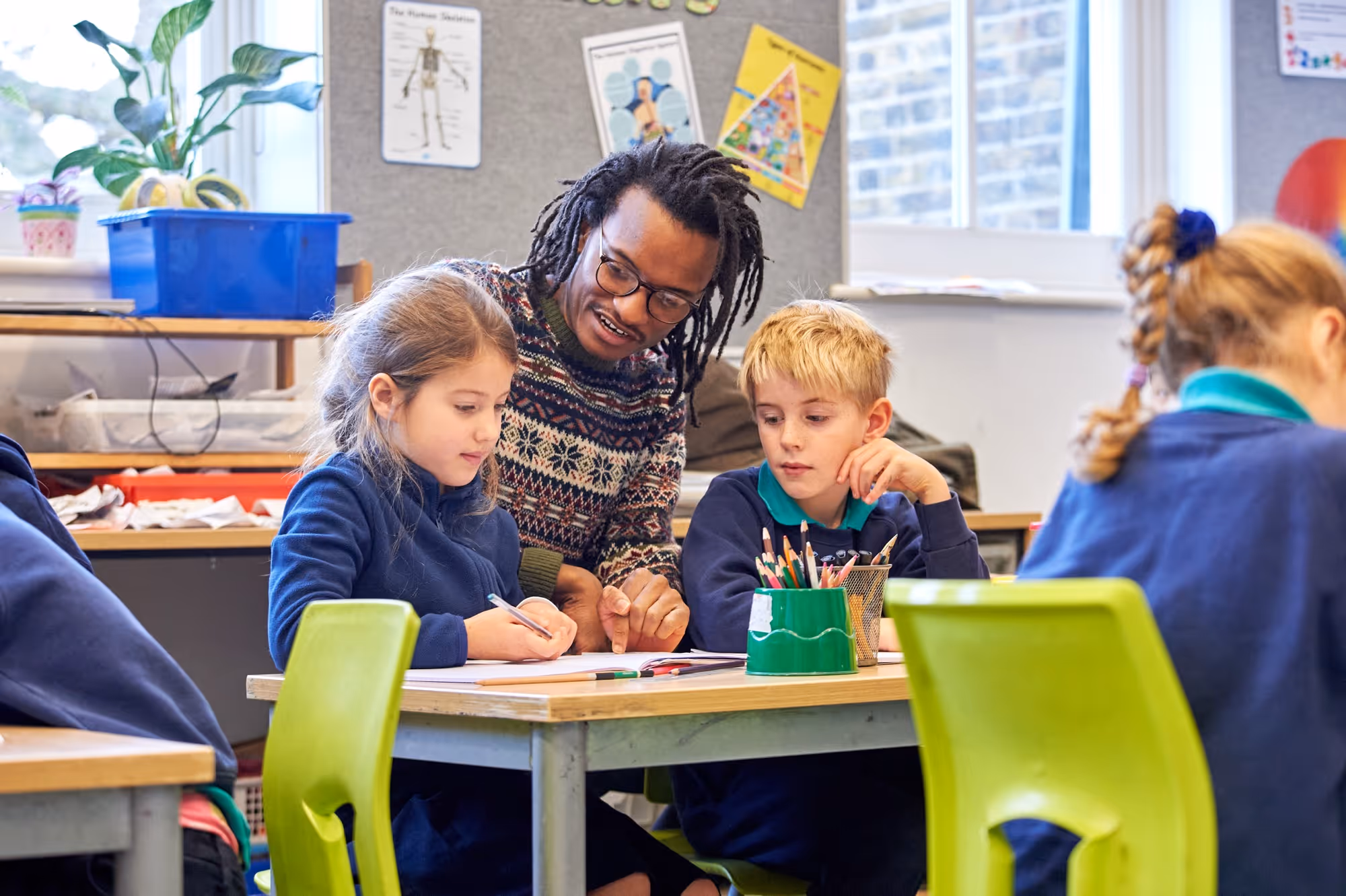 A male teacher with glasses and dreadlocks assists two young students, a girl and a boy, sitting at a classroom desk with pencils, paper, and a green pencil holder, while another student works nearby.